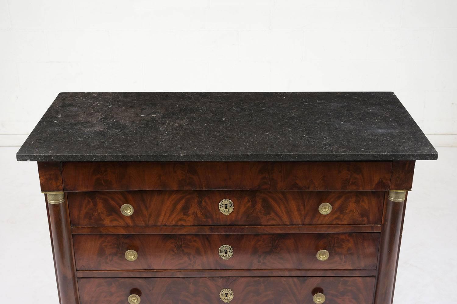 This 1840s French Empire-style commode is covered in Flemish mahogany veneers with the original finish. There are four drawers with the bottom three adorned with decorative bronze pull knobs and keyhole plates. Flanking the drawers are carved