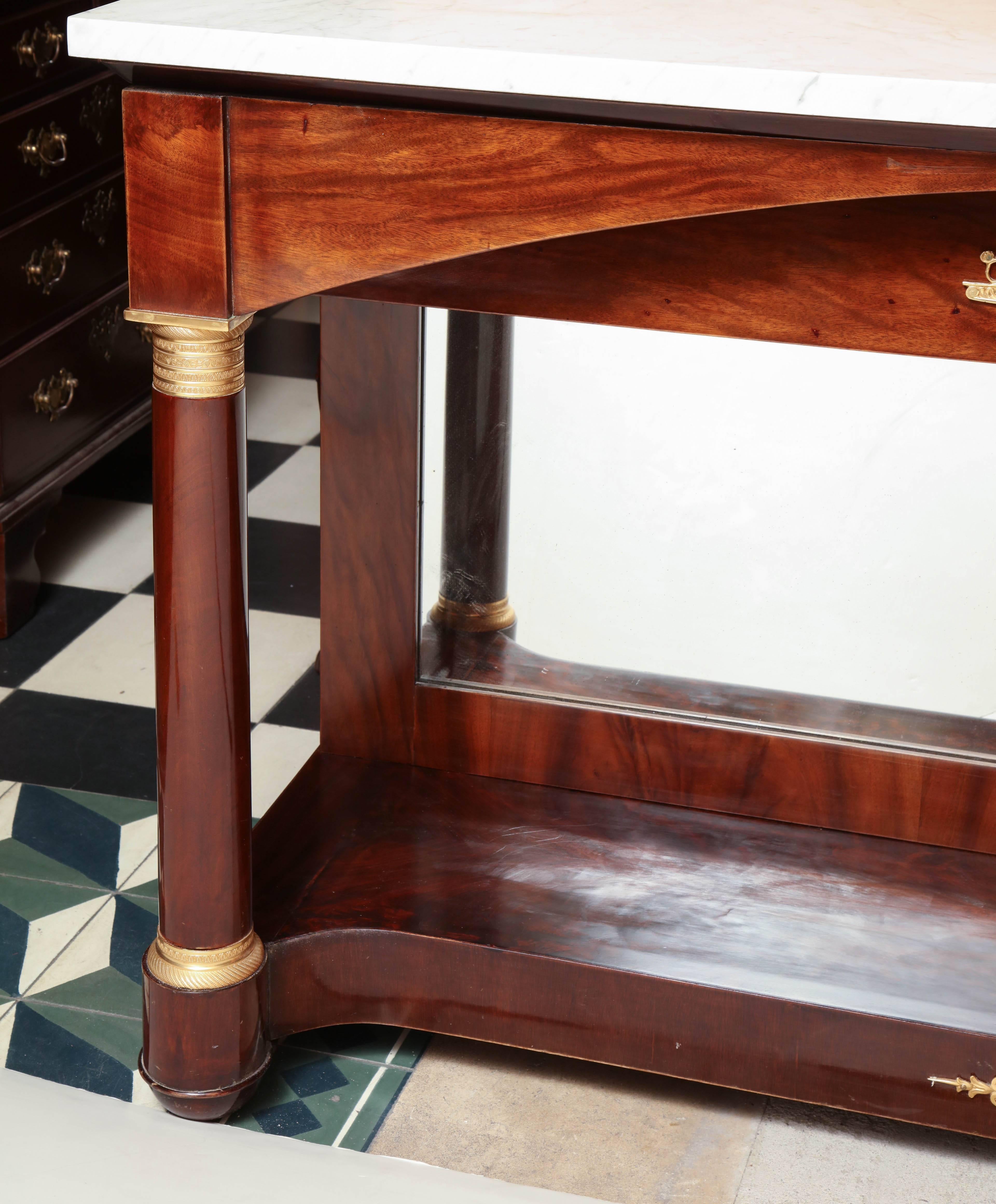 Long French Empire mahogany console table with Carrara marble top, bronze mounts, mirror back and bronze capital column supports with a shelf stretcher base.