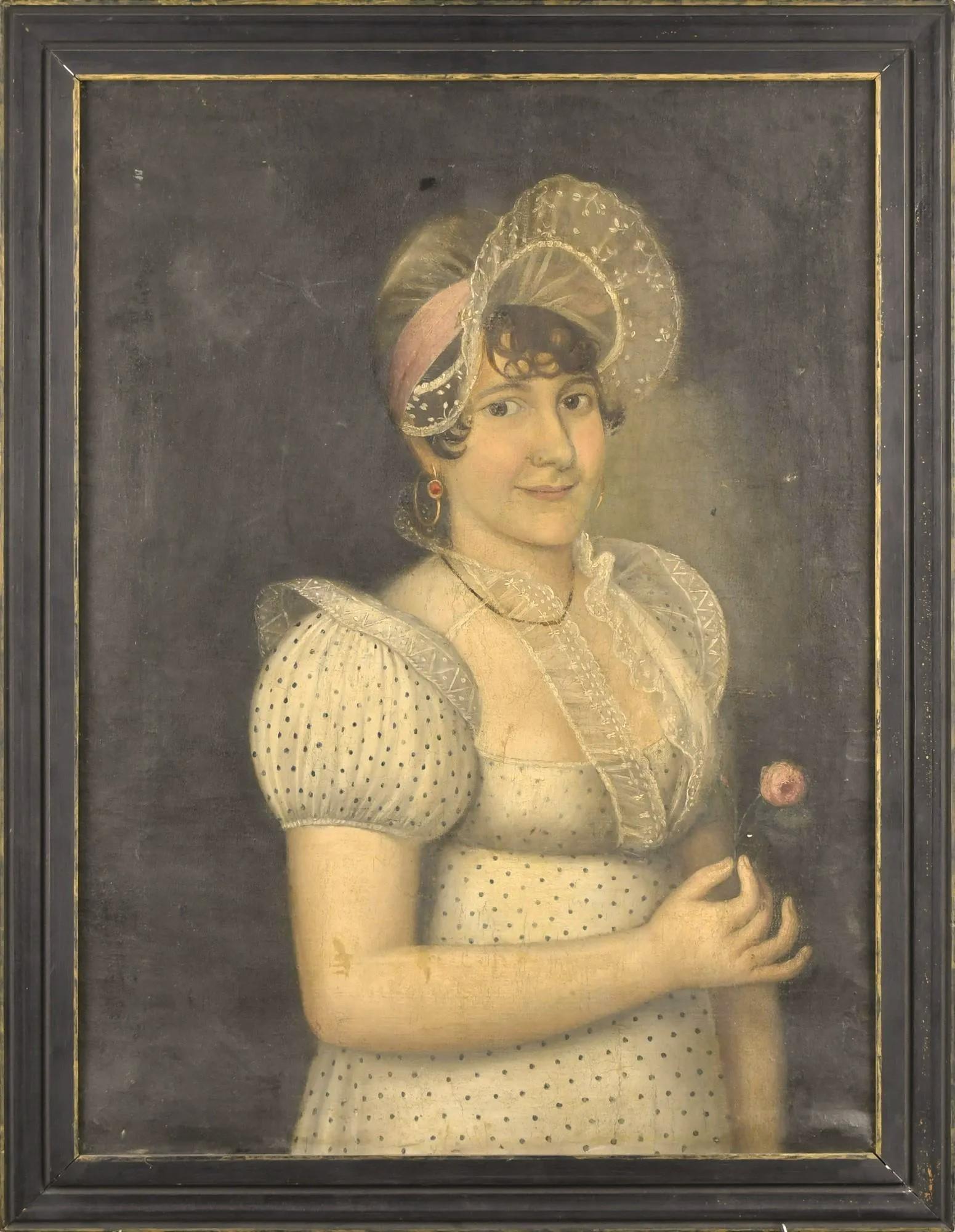 An early 19th century naive oil-on-canvas portrait of a young woman dressed in a laced trimmed dress and holding a flower, Empire period. The reverse showing a French gallery label. Older black wood frame.