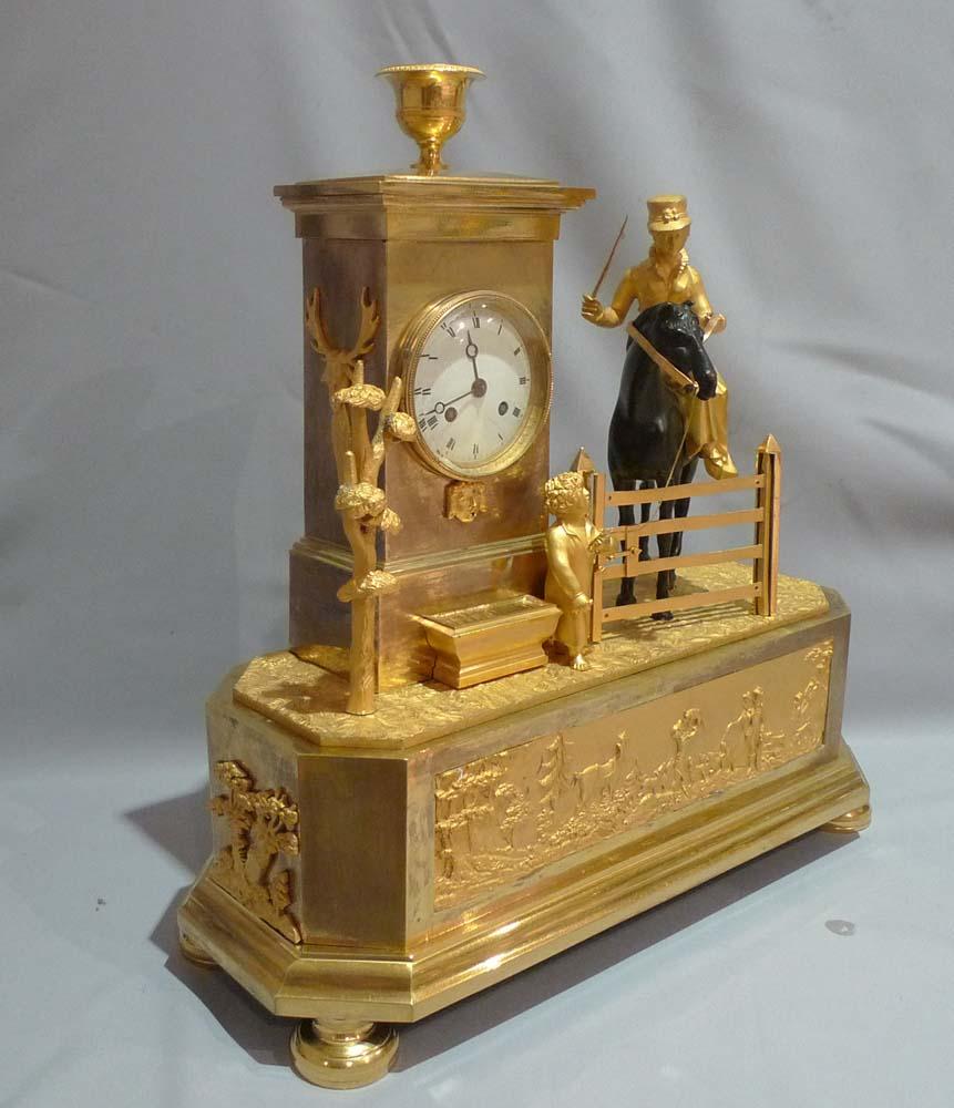 A very unusual French Empire Genre Arcadian mantel clock of a hunting scene. The primary subject is that of a finely dressed woman in riding habit, hat, whip and gloves riding side saddle on a fine patinated bronze hunter. A young boy is leading the