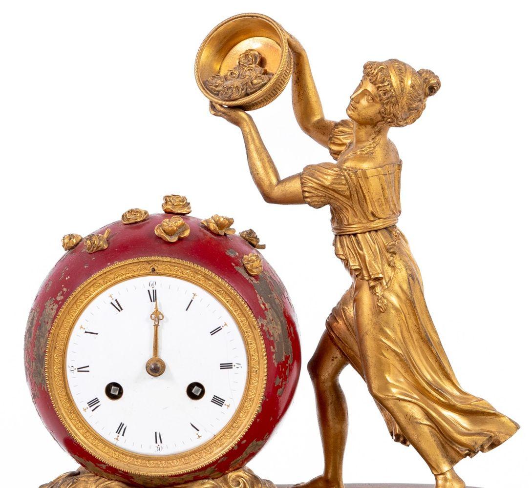 French Empire Gilt Bronze Figural Clock, circa 1830, the classical muse upholding a basket of flowers beside a red-painted globular clock with white enamel dial, the movement strike on bell, on an oval base on four feet. 14