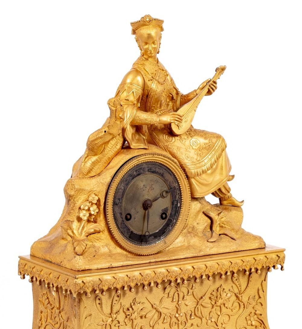 French Empire Gilt Metal Figural Clock, circa 1840, in two parts, the upper section with a seated lute player aside the circular metal dial, the movement strike on bell, the lower section richly decorated with foliate scrolls on a scrolled base.