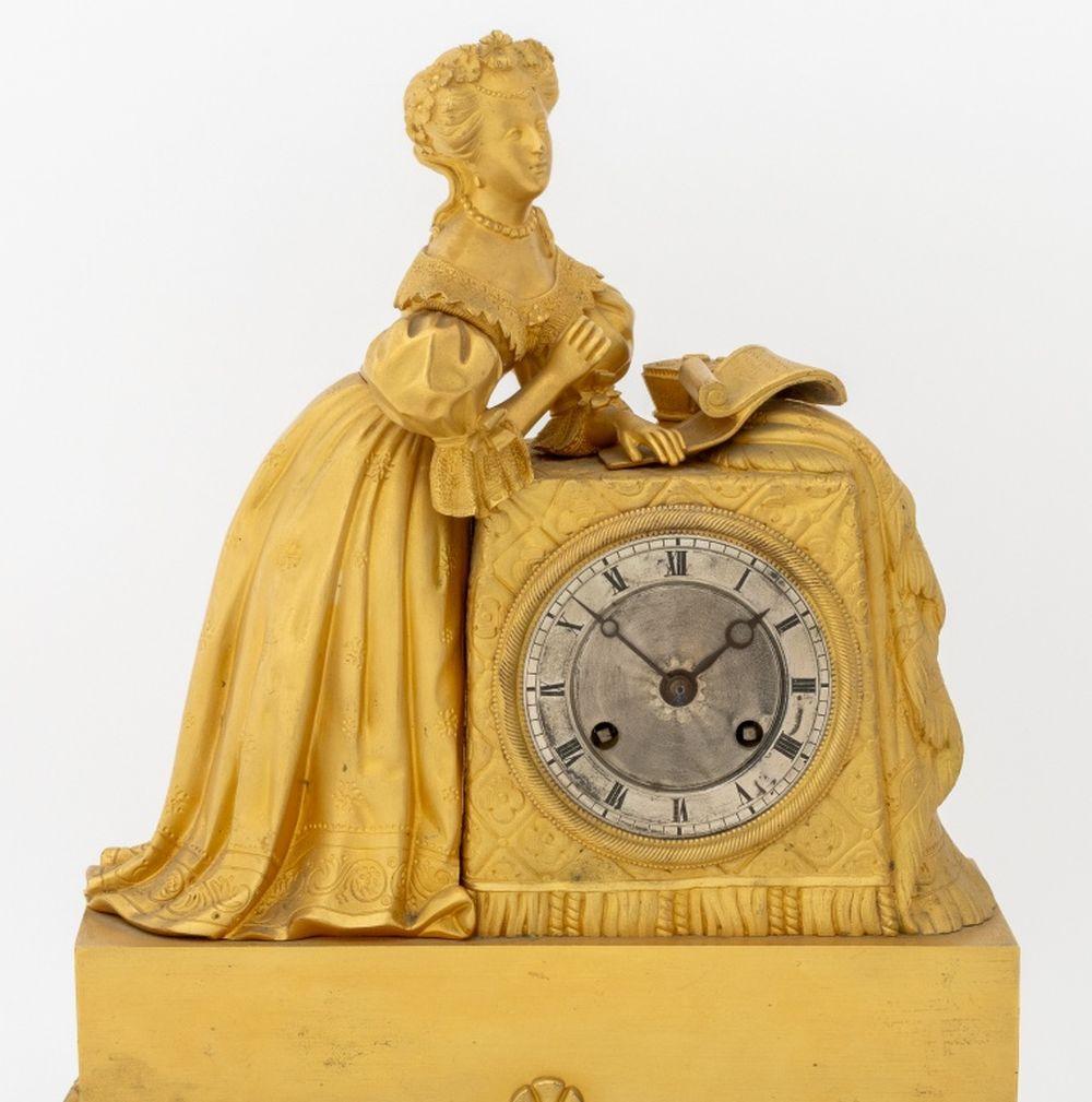 Pendule figurative en métal doré de style Empire français, vers 1830, le cadran circulaire avec mouvement de sonnerie sur cloche, surmonté d'une femme écrivant, sur une base à acanthes et cannelures avec des pieds à volutes. 17.5