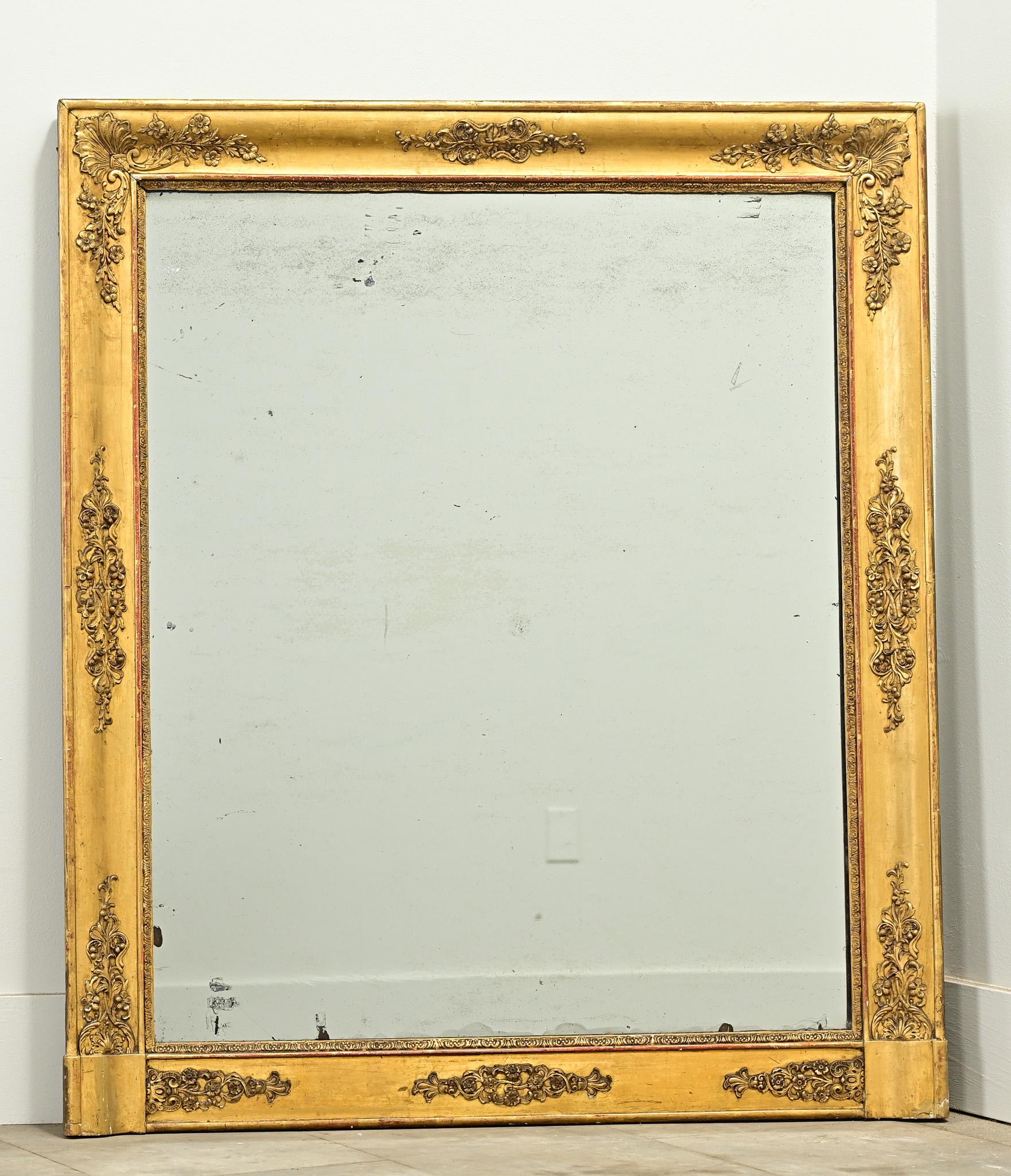 A French Empire gold gilt mantle mirror from the 1800’s. The carved frame has its original gold gilt finish revealing the reddish bole beneath with decorative floral applique details. The original mirror plate has foxing and light crystallization.