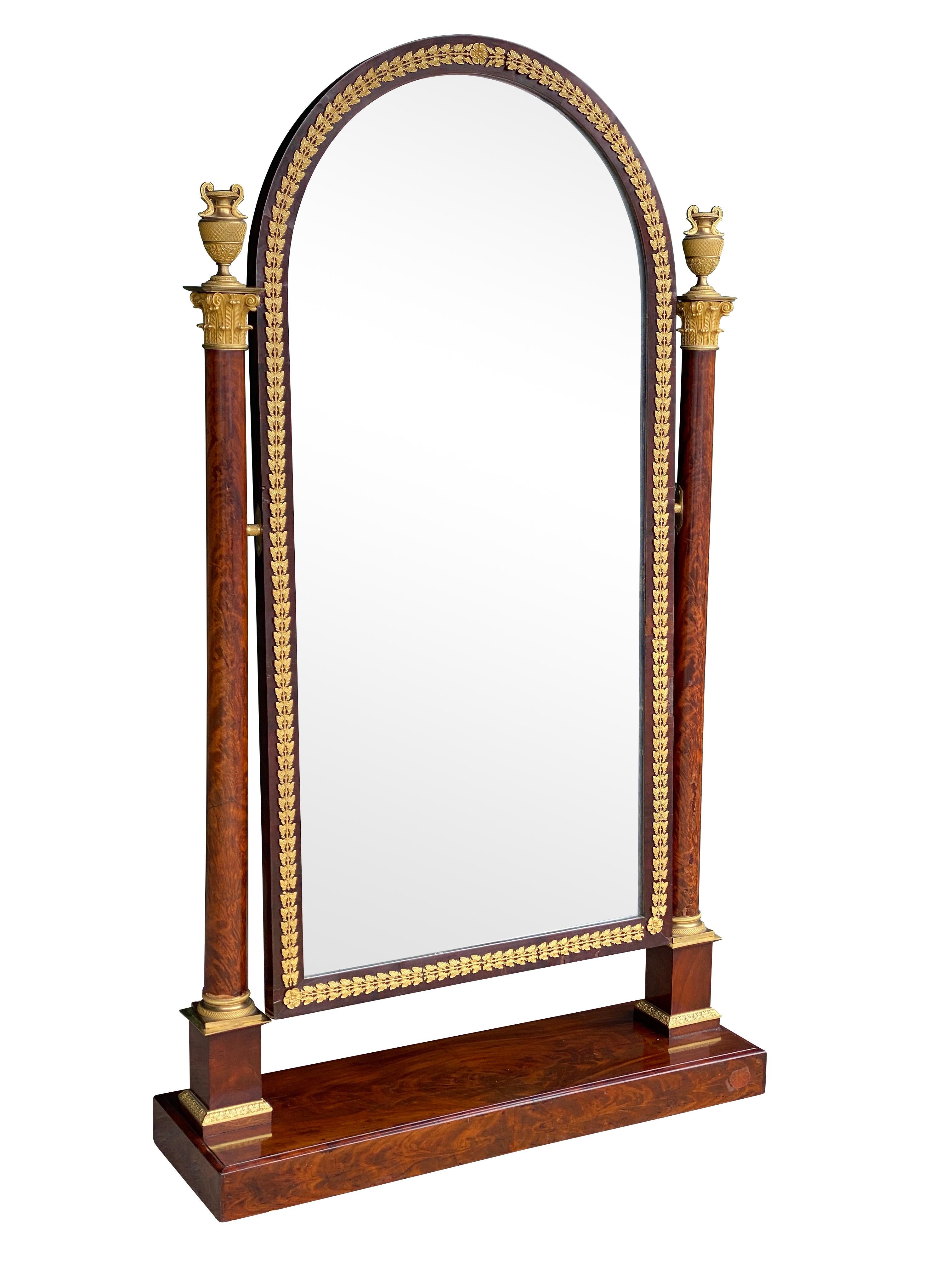 With an arched mirror set in a conforming frame and able to swing and adjust, flanked by a pair of columns with Corinthian capitals and urn finials, square plinth base. Casters.