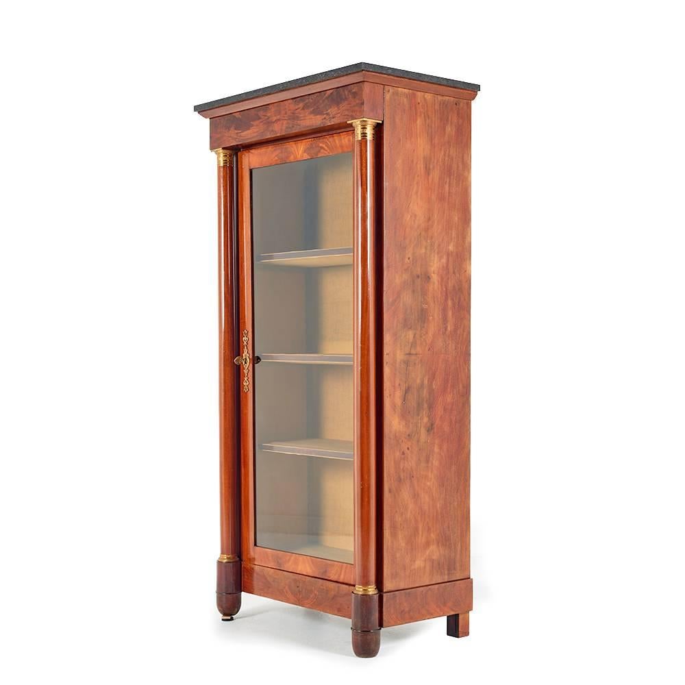 An unusually-narrow French Empire style single-door bookcase or cabinet, the case veneered in figured flame mahogany, the single door flanked with columns, each with brass bass and capital.
The piece is capped with a dark grey marble top, and also