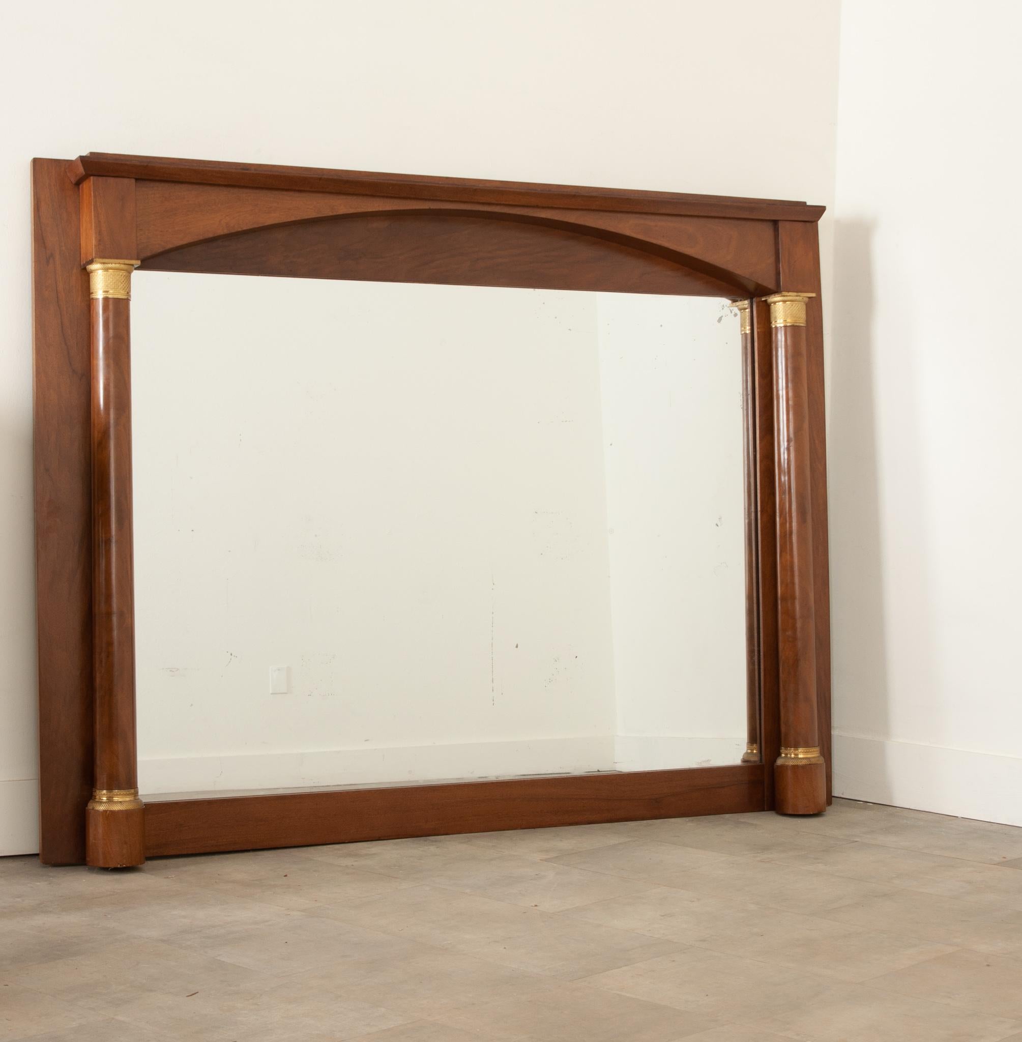 A stately, Empire style mantle mirror from France with some light foxing throughout the original mercury mirror plate. The minimally carved mahogany has gained a fine patina. Bright, cast brass capital and bases adorn the column forms flanking the