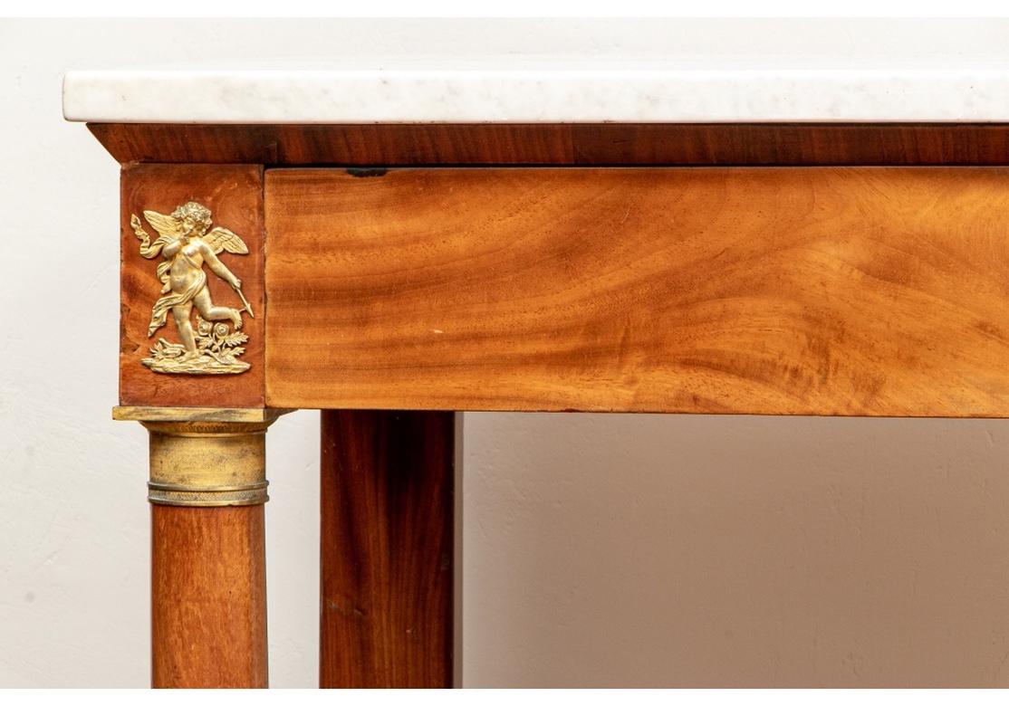 A French Empire white marble top console with a central bronze escutcheon comprised of two nudes with scrolling feathered tails; flanked by putti motifs, above columns and the whole resting on a plinth base.
Dimensions: 57 1/2