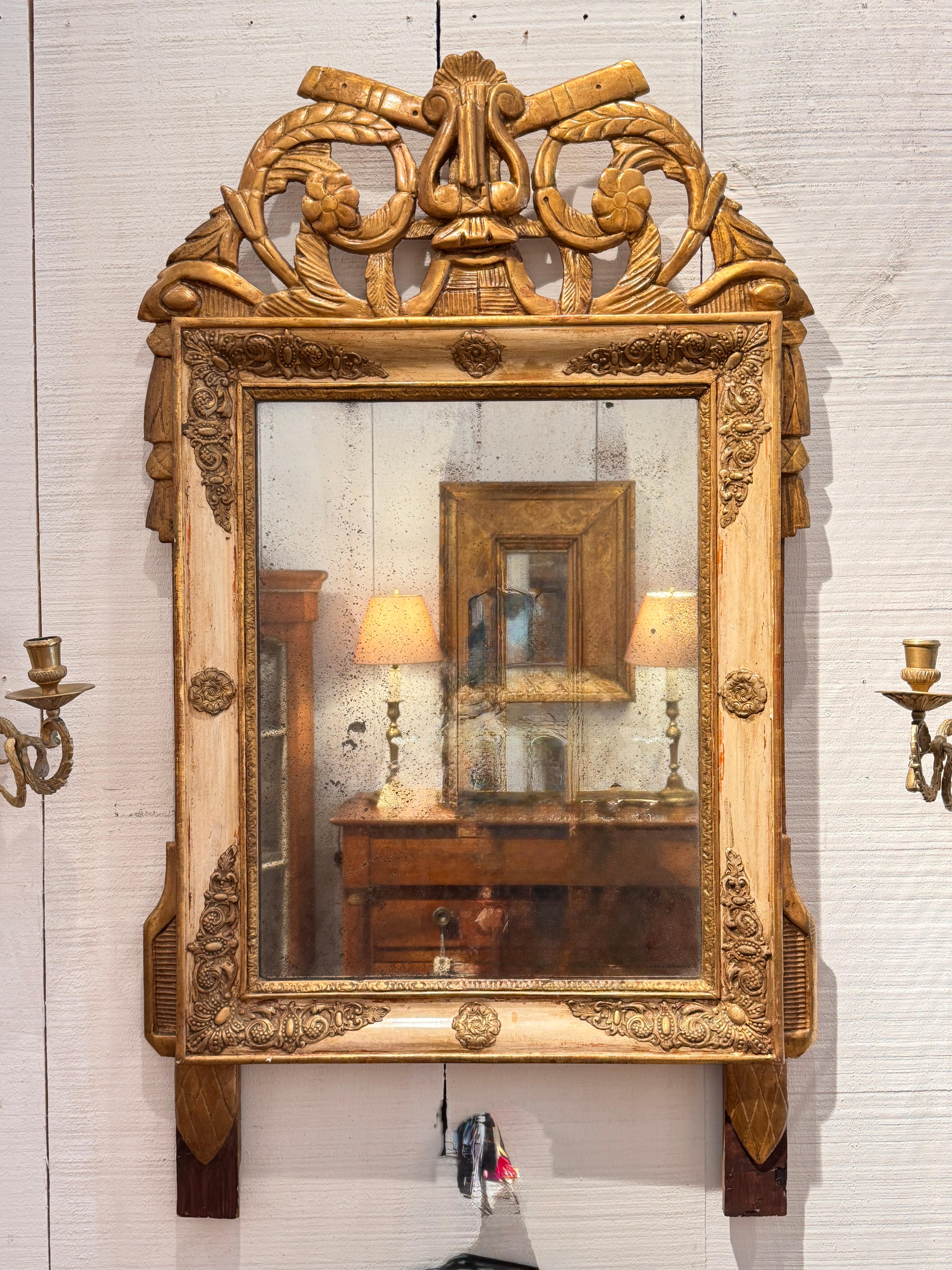 Carved and gilded French Empire mirror featuring lyre and scrollwork at the crest, with decorative rosettes and corner elements. The aged mirror plate adds character and period charm.