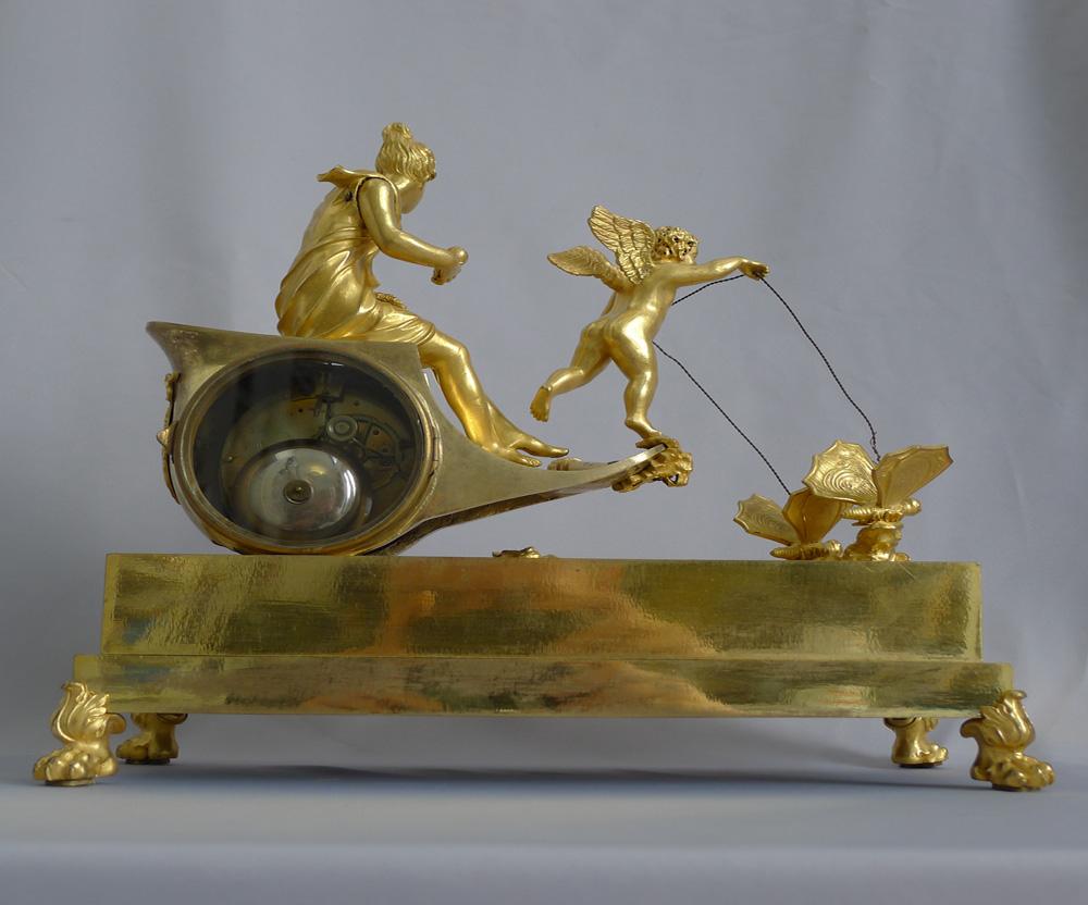 Pendule antique Empire en bronze doré signée sur le cadran en émail annulaire Ravrio Bronzier. Cupidon contrôle le char tiré par une paire de papillons, tandis que Vénus est assise dans son char, tenant un cœur dans une main et une couronne dans