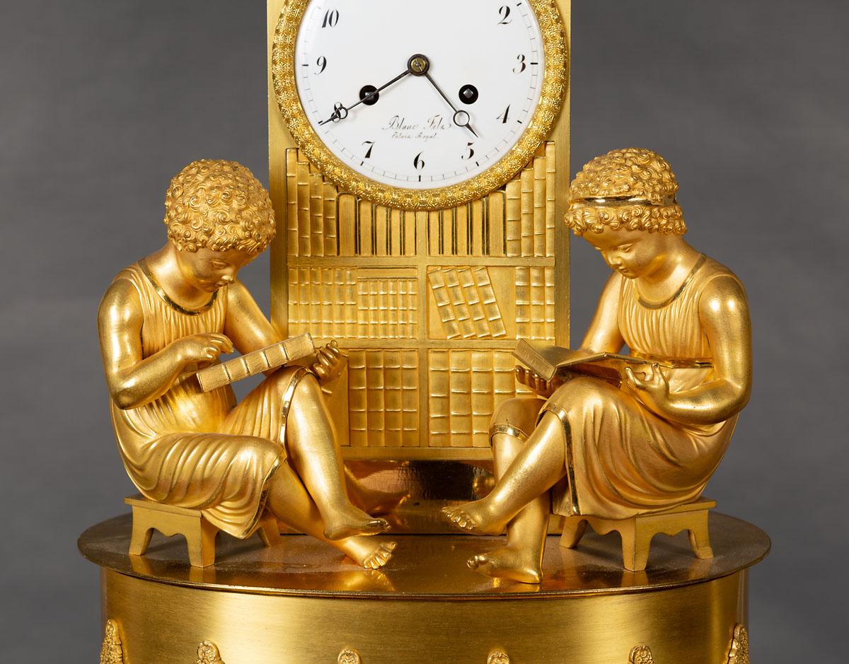 French Empire Ormolu mantel clock resting on four turned feet supporting an oval base with raised garland and zig-zag rope decoration. Two scholars are seated upon the base both studying books in front of the ‘library’ below the clock dial. The two