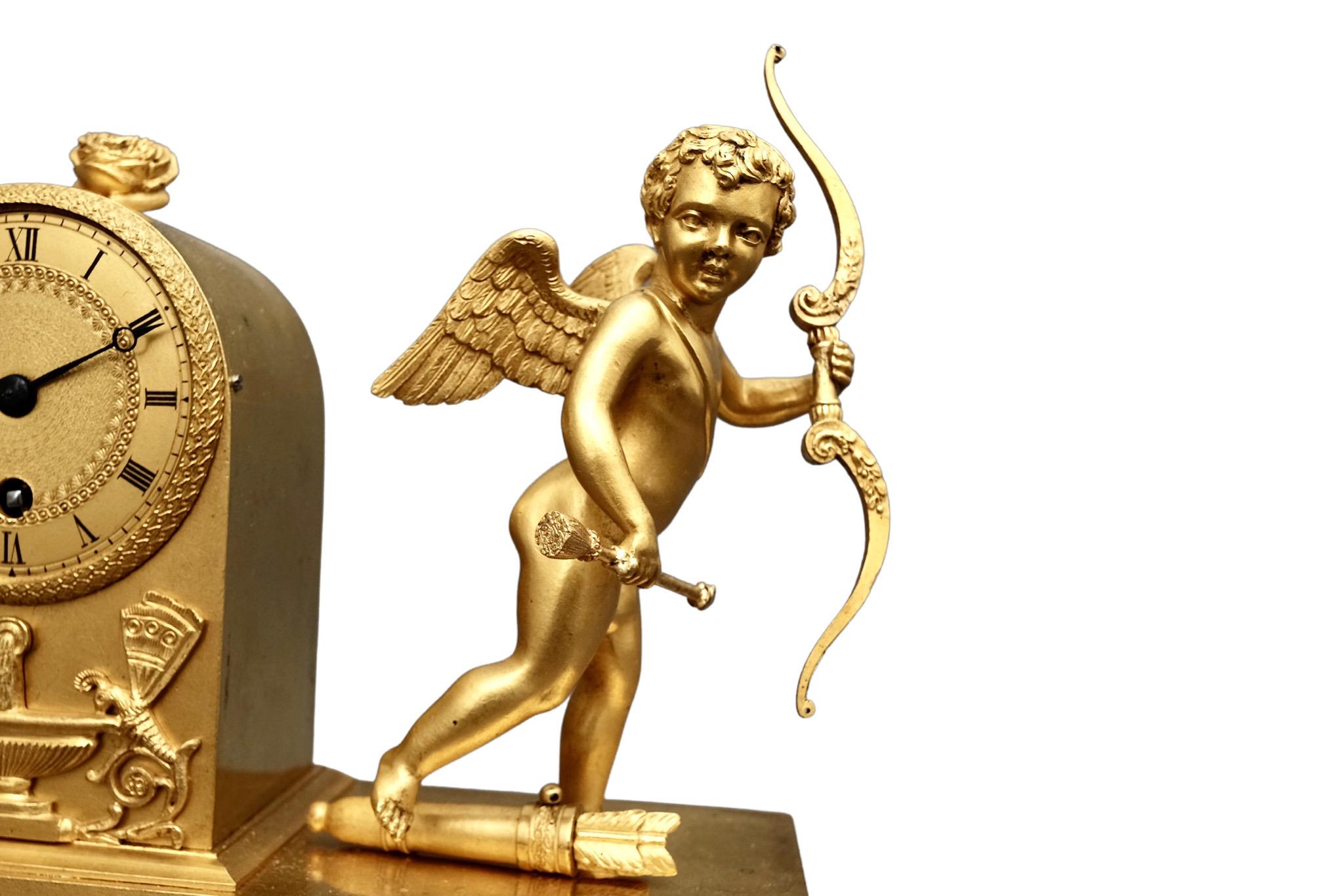 A delightful French Empire miniature ormolu timepiece mantel clock featuring a cherub with wings, bow and arrow. The cherub has one foot resting on a quiver holding more arrows and stands alongside the beautifully decorated dial with Roman numerals