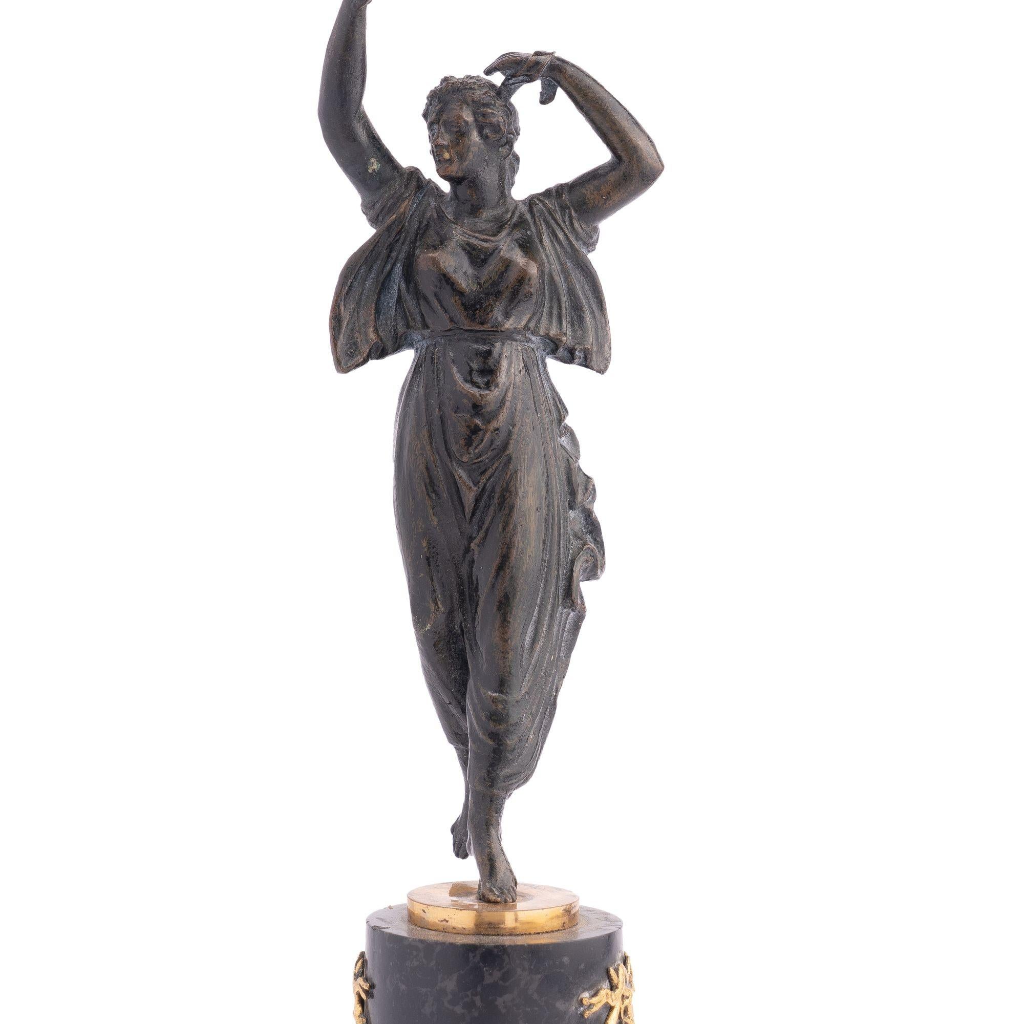 Patinated and parcel gilt cast bronze figural candlestick on a circular marble base with square plinth. The figure, standing on one foot, holds a leafy form fire gilt candle cup above her head.
France, Empire Period, circa 1800-10.