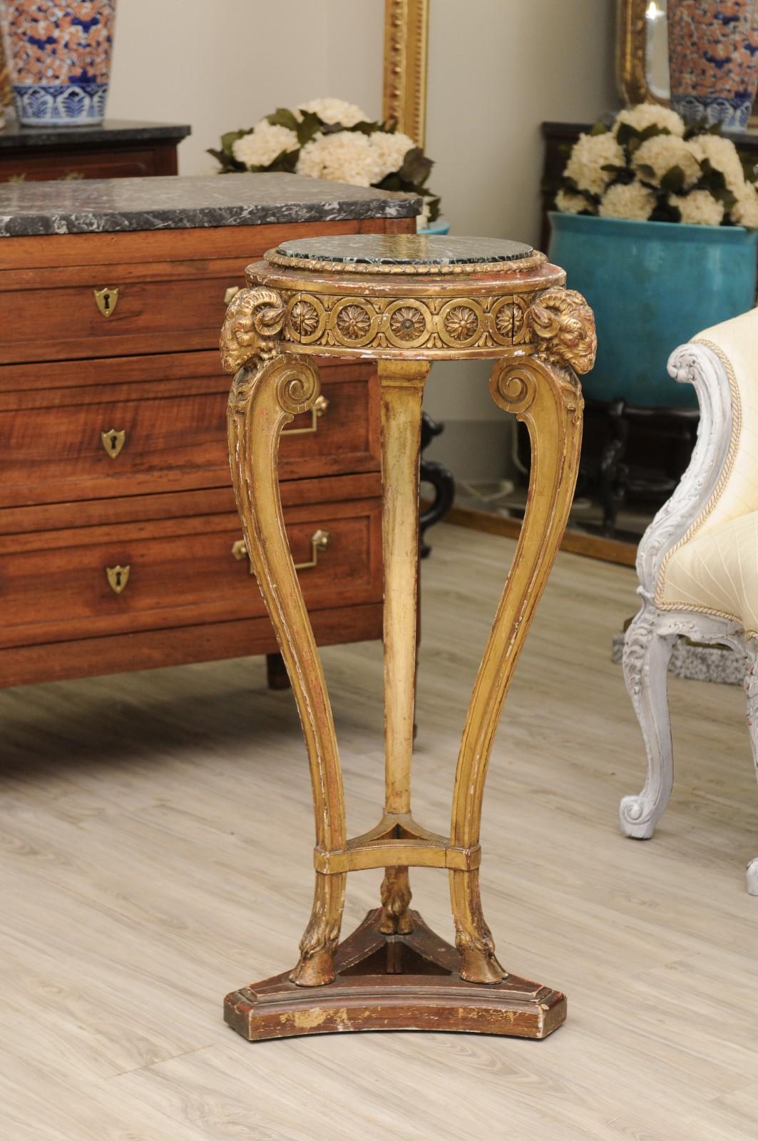 This highly carved period Empire pedestal has carved rams heads on three corners which are at the top of the 3 curved legs with paw feet. The legs stand on a wooden triangular shape base. Giltwood and original marble top.