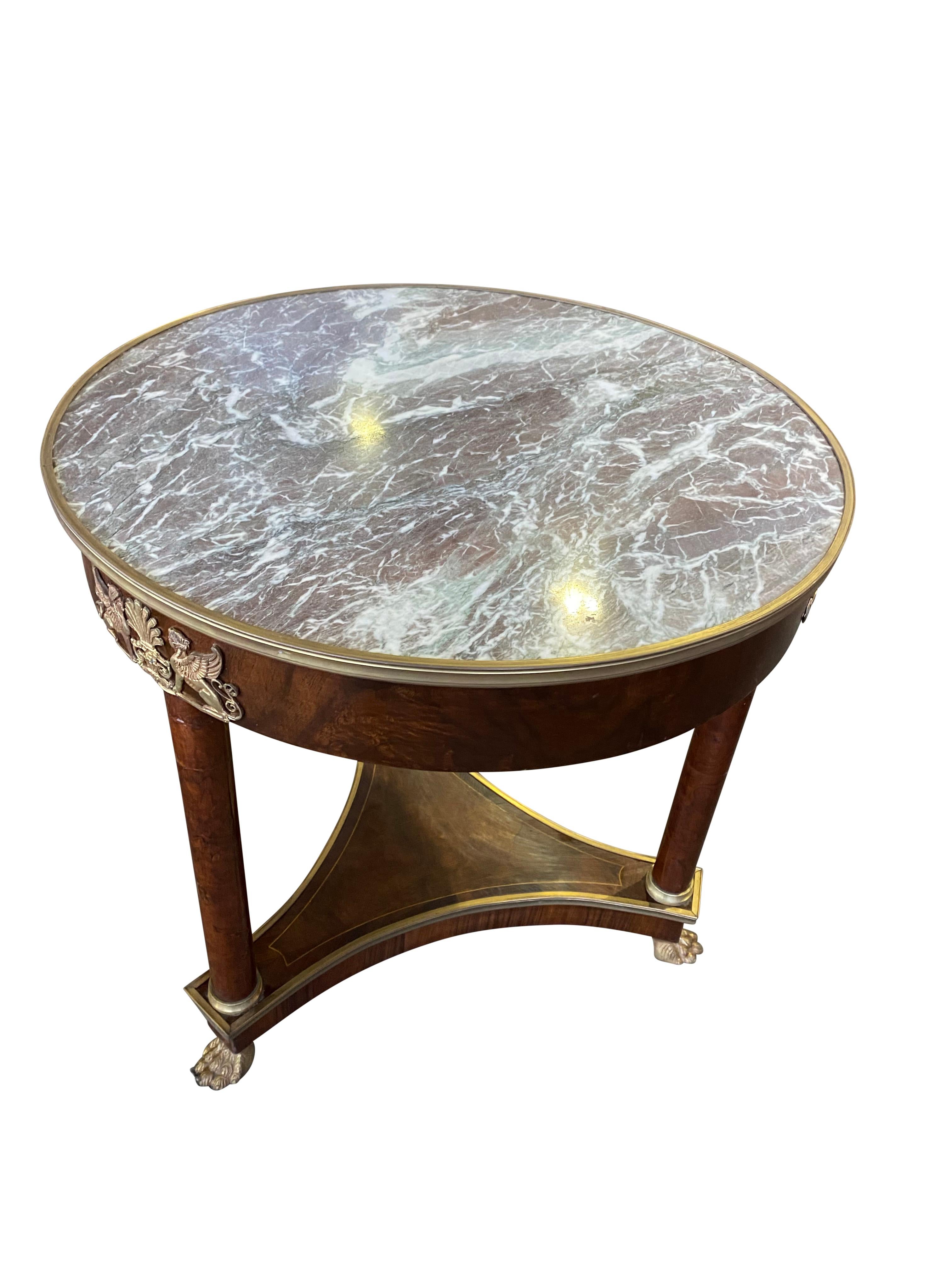 A fine 19th century French Empire round marble-top mahogany table with three legs separated by a triangular stretcher. The unique curved trip base supporting the table is lifted by golden paw-like feet beneath the legs. 

Dimensions (cm)
80 H/80