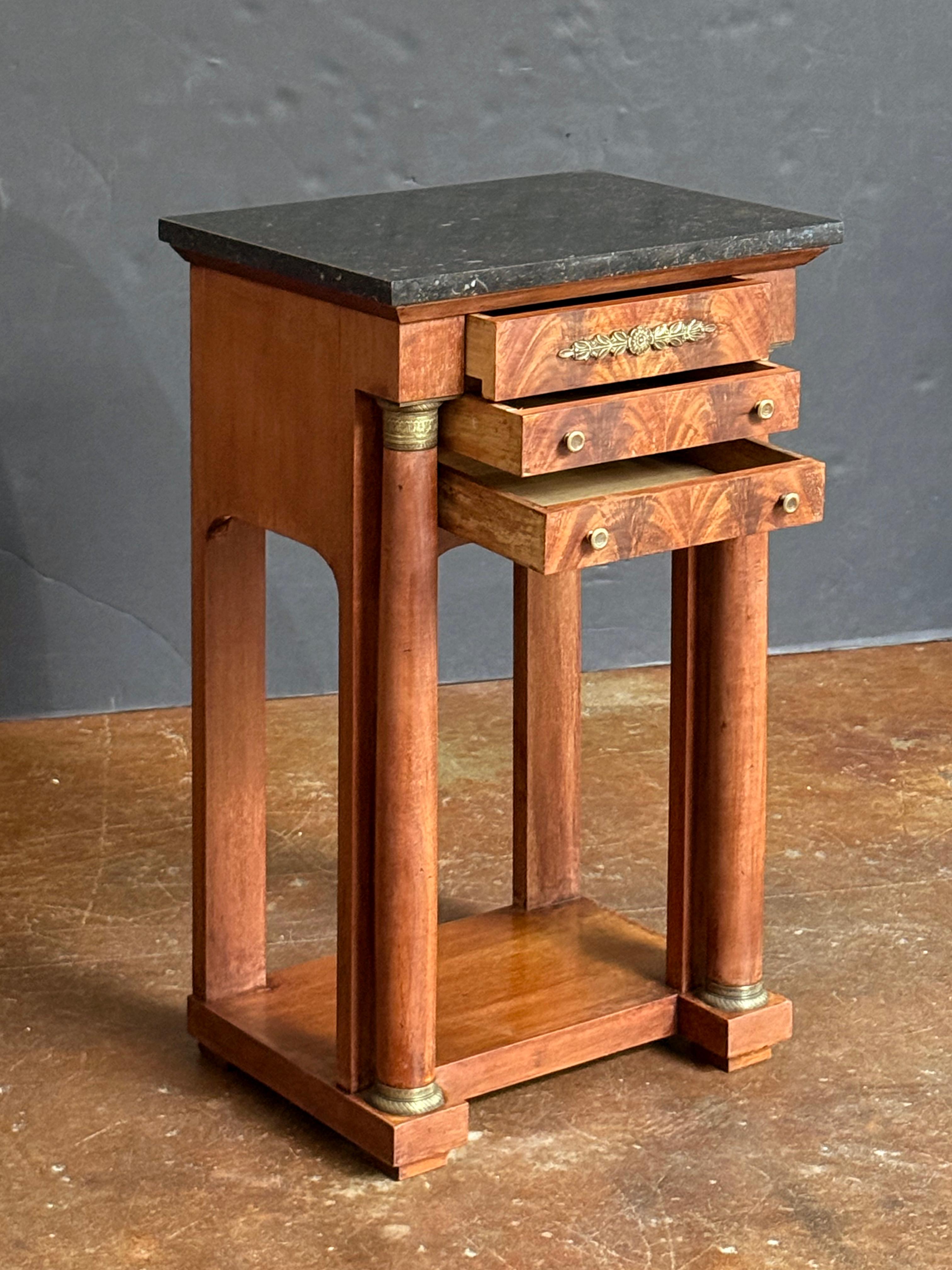 Bellissimo tavolino o comodino di epoca Impero francese in mogano tagliato a fiamma con piano in marmo nero figurato, caratterizzato da un fregio con un cassetto incastrato con placca decorativa in ormolu, sopra due cassetti incassati, con