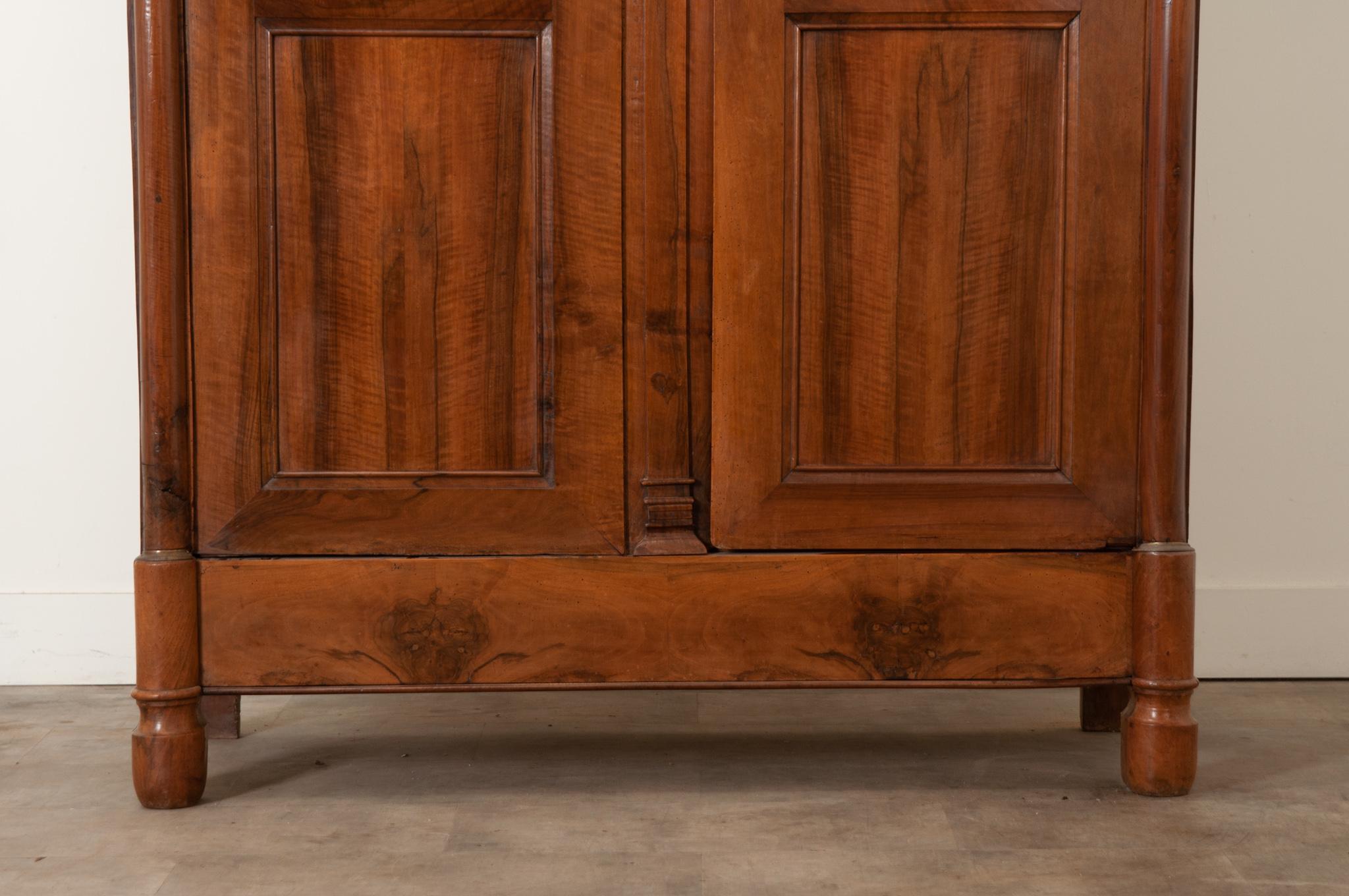 A handsome solid walnut Empire armoire, made in France during the 19th Century. This armoire features a pair of large bookmatched and paneled doors on hidden hinges, flanked by columns capped with gilt bronze ormolu at its capitals and bases. The