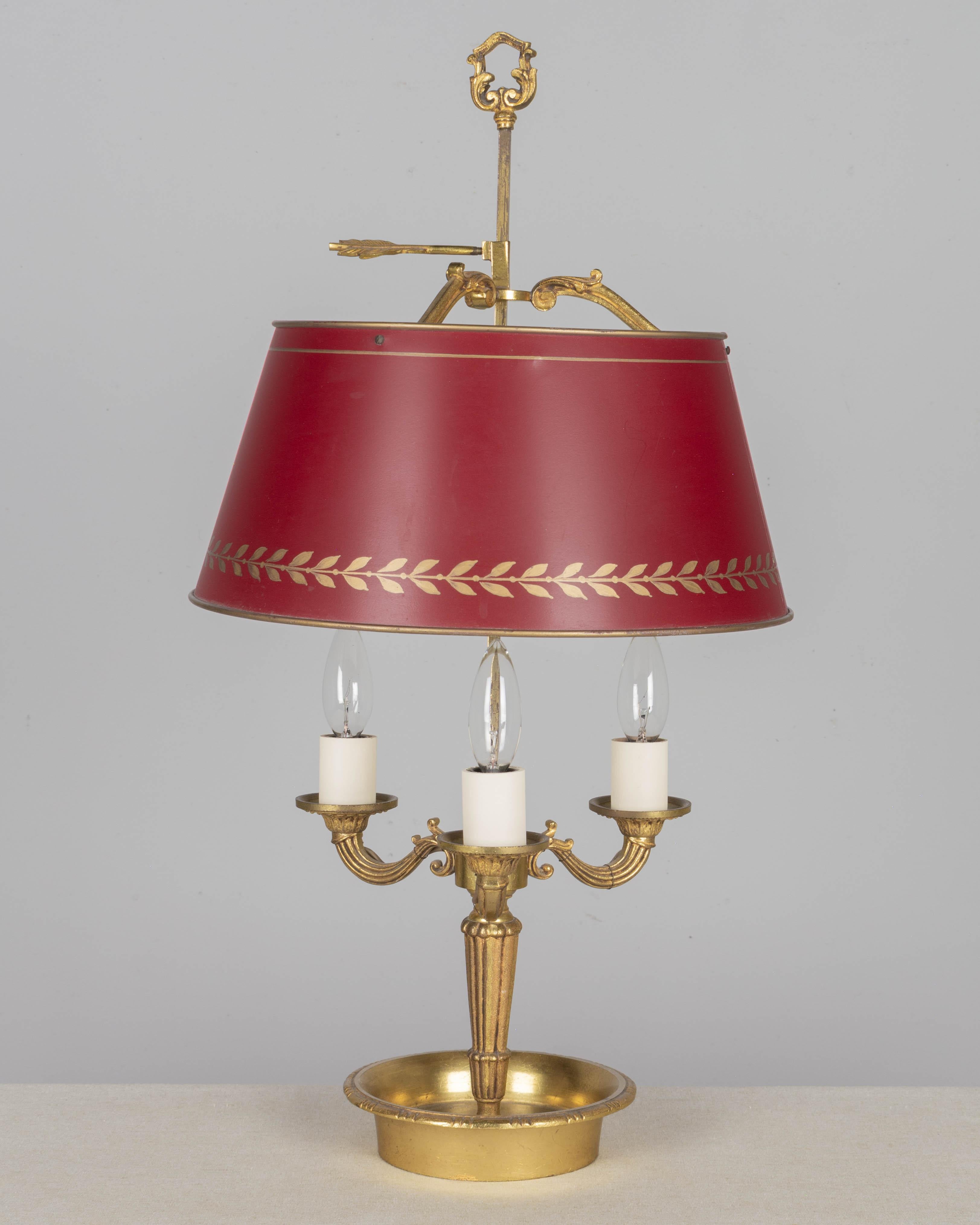 Lampe de table bouillotte de style Empire français avec bougeoirs décoratifs en laiton moulé à trois lumières et abat-jour en tôle rouge réglable en hauteur. De nouvelles couvertures de bougies. Câblé et en état de marche. Vers 1970. 
Dimensions :