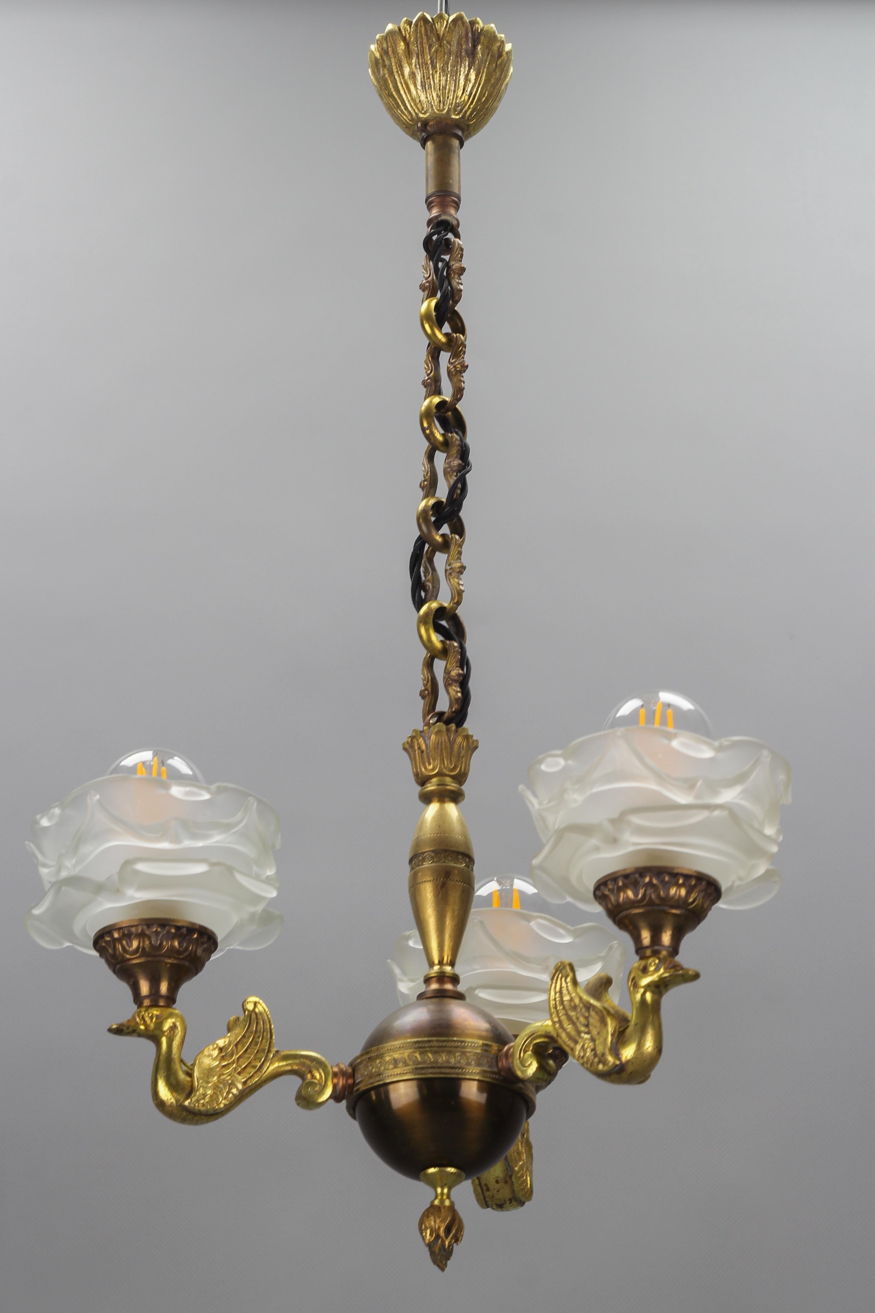 French Empire style brass, bronze, and frosted glass three-light chandelier from ca 1950s.
This adorable and compact Empire-style chandelier features a circular brass body and three arms - three bronze swans each holding a frosted glass lampshade