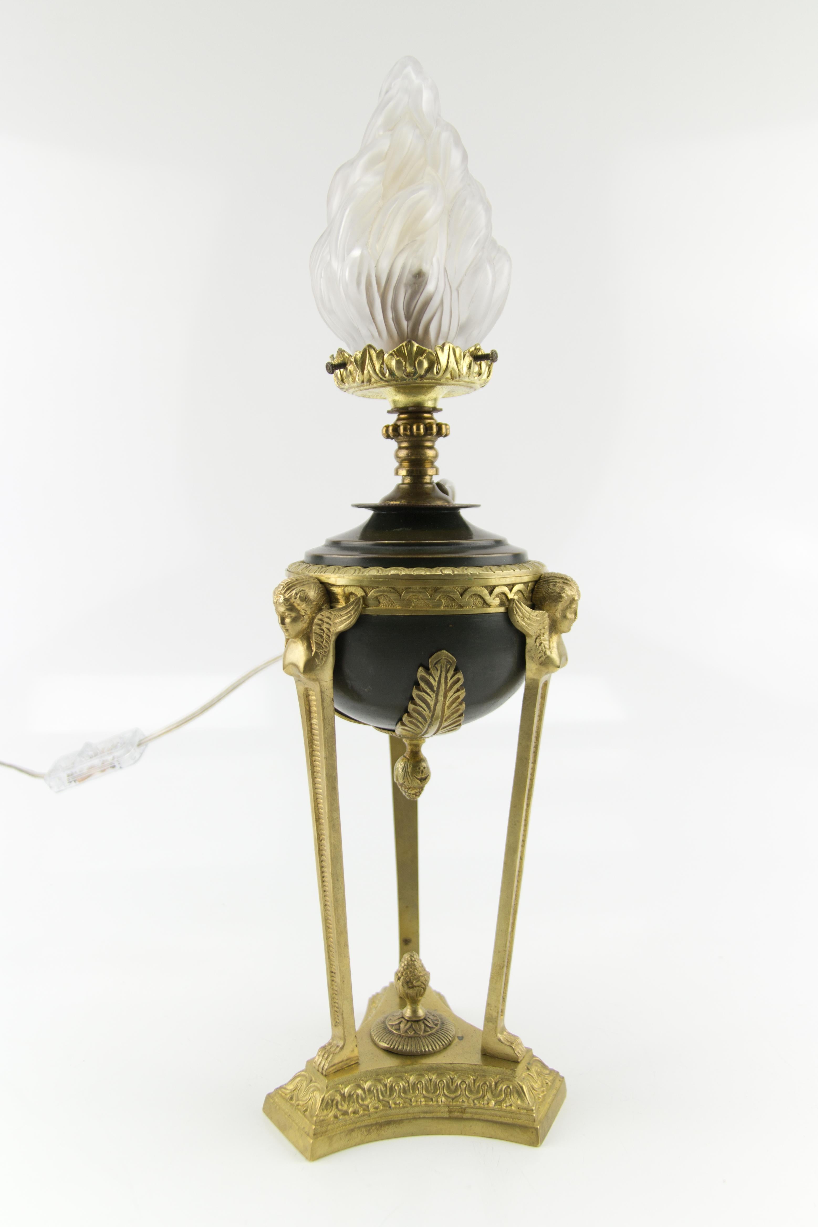 Early 20th French Empire style table lamp has a tri-form bronze plinth with a central pine cone, with three legs with paw feet topped by three bronze sphinx supports, holding the dark green tôle oil reservoir, decorated on all three sides by bronze