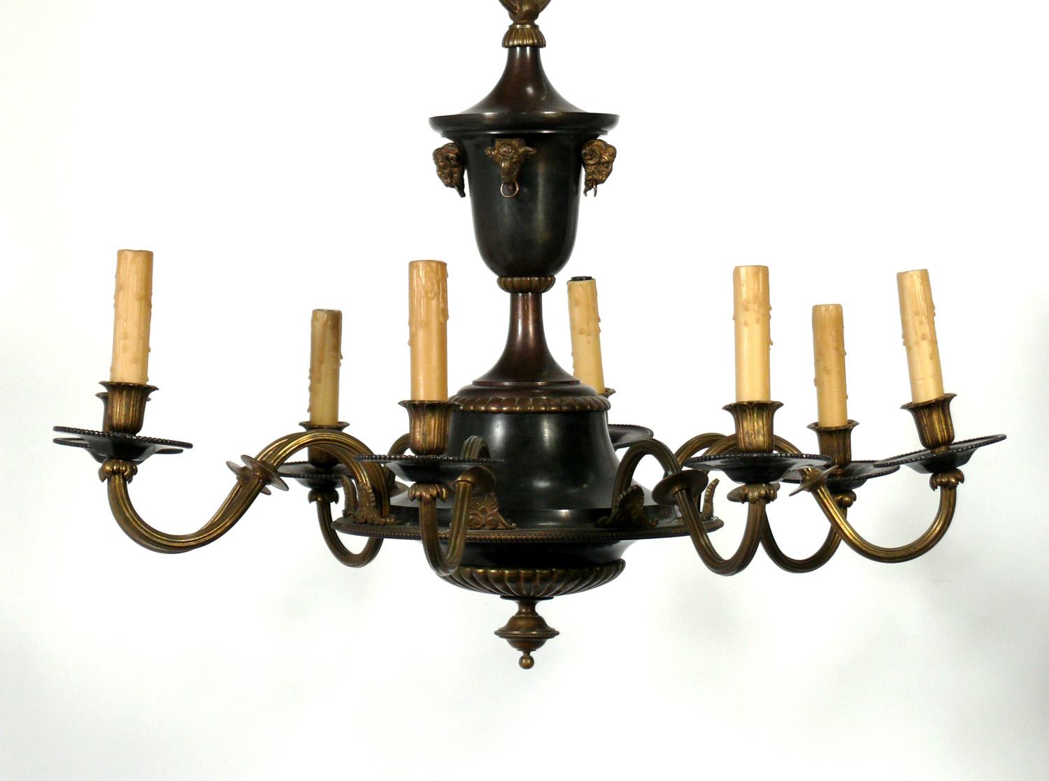 French Empire style eight arm bronze chandelier with ram’s head details, France, circa 1930s. Rewired and ready to mount. This eight light chandelier has scroll and acanthus leaf details at each fluted and curved arm. The bobeches have a faceted