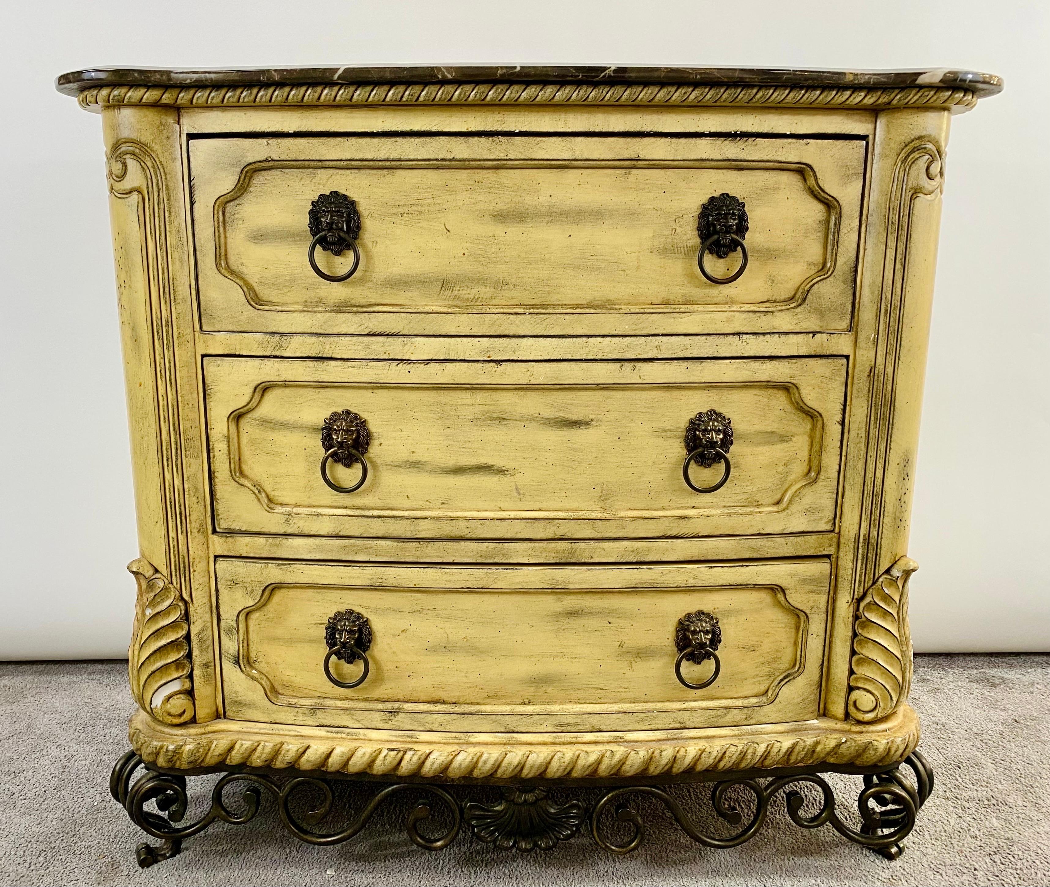 An exquisite French Empire style commode or dresser offering three drawers. The commode or dresser is painted in antiqued beige tone and features lion head bronze pulls. The dresser or commode is raised by finely carved tole metal bottom showing