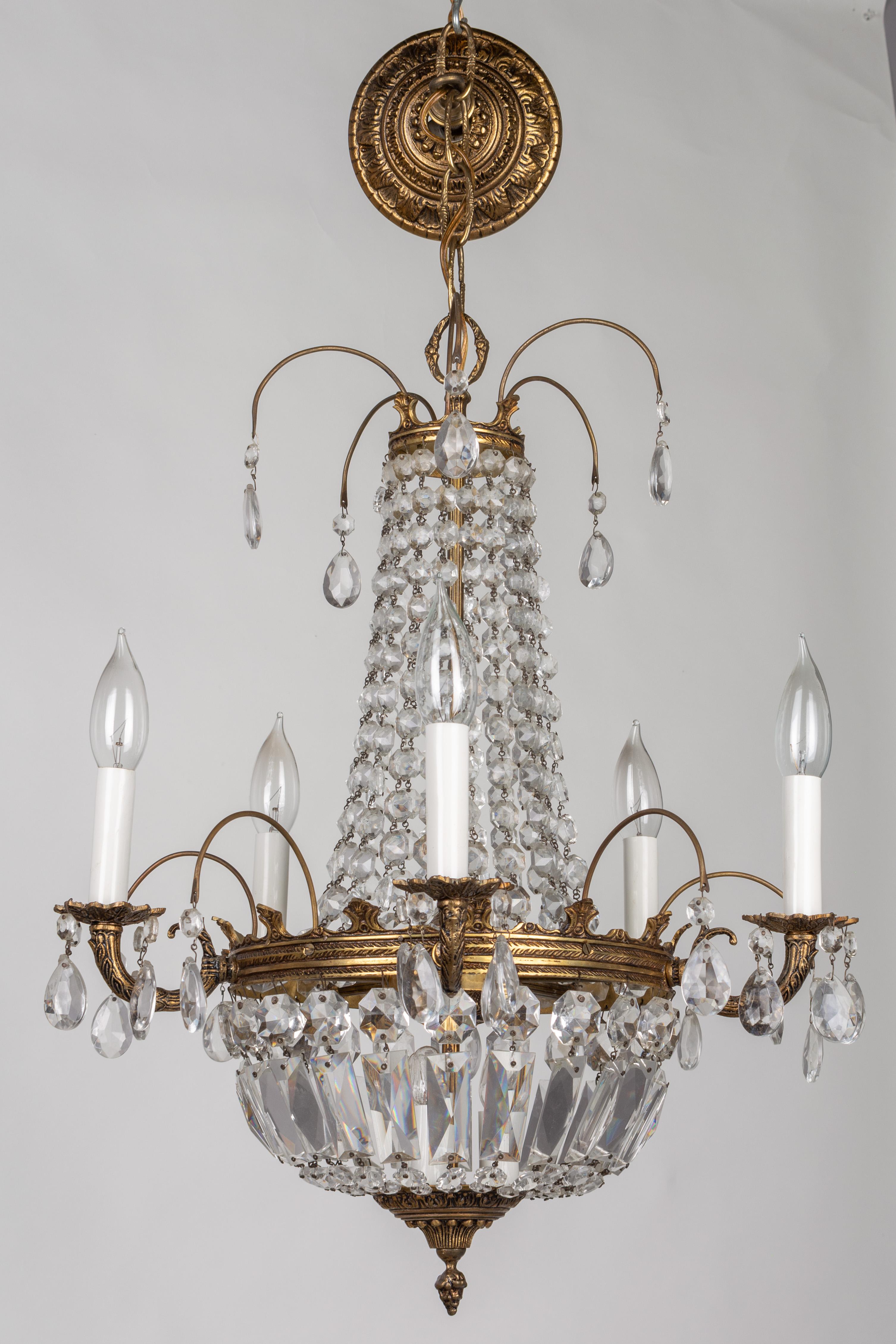 A French Empire style crystal chandelier with six brass candle branches and three interior lights. Graduated crystal prisms drape down to central embossed brass ring. Strands of octagonal crystal jewels and baguettes form the basket below. Original