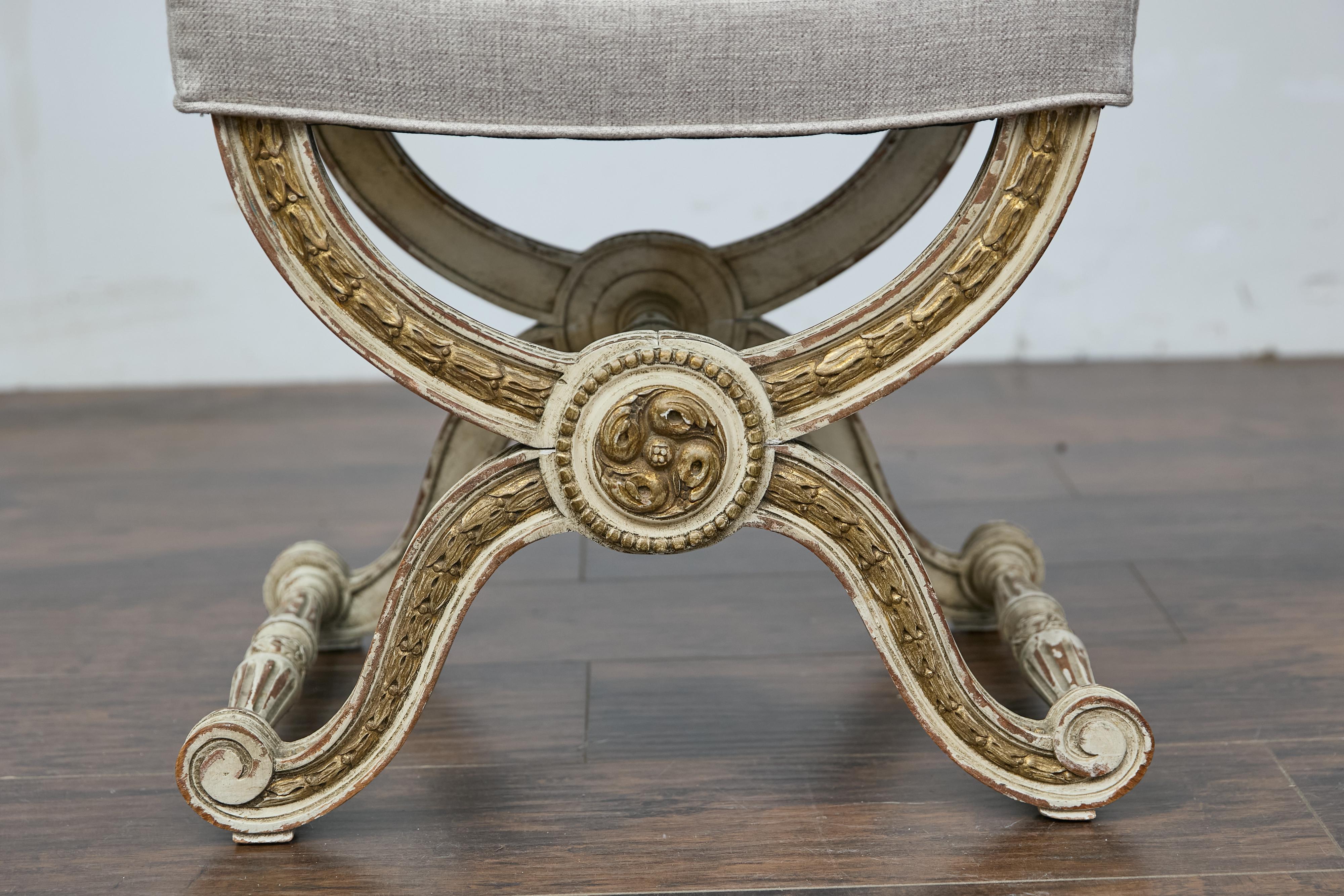 Sgabello a curule in legno dipinto in stile Impero francese del 1900 circa con accenti dorati, gambe a X ricurve, motivi a campanula e rosette intagliate, appena rivestito in tessuto di lino. Questo elegante sgabello in stile Eleg, realizzato