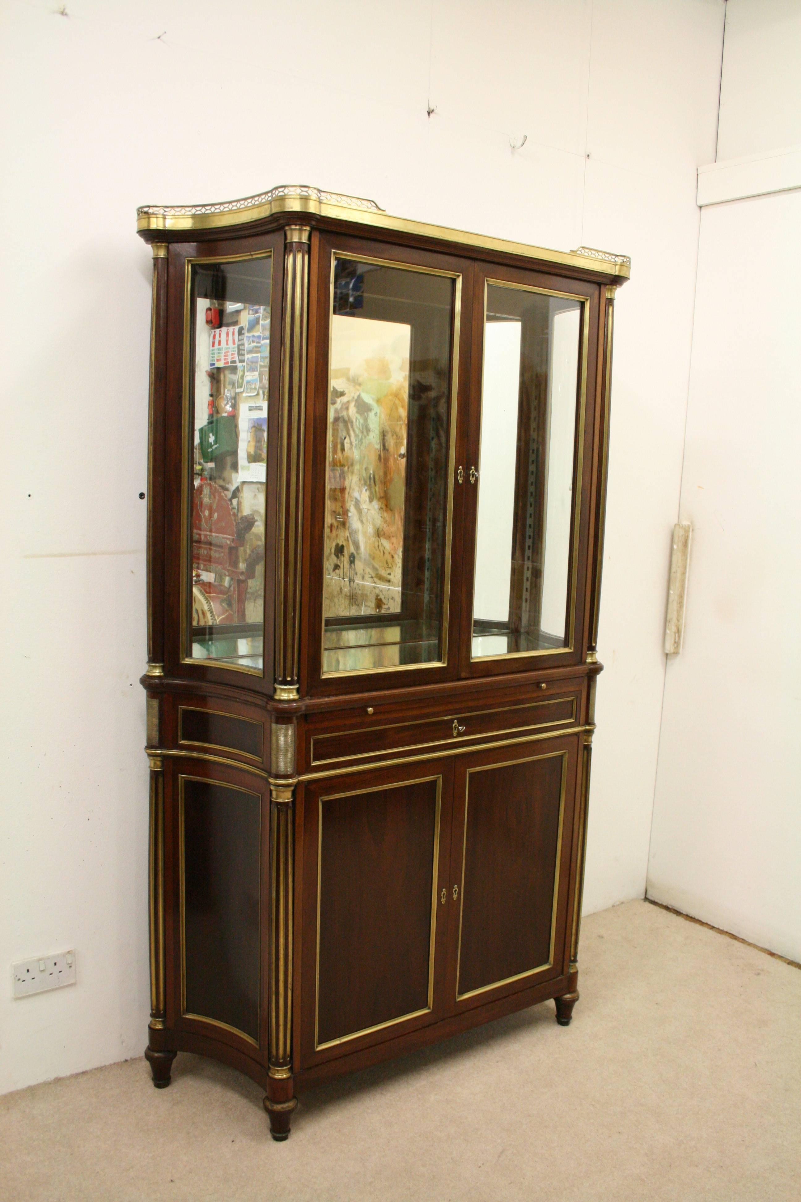 French Empire style mahogany and brass inlaid display cabinet or bookcase with a brass fretted gallery above the glazed doors and fluted brass inlaid pilasters, circa 1900. Inside there are shaped mirror shelves and a mirror back. The base section