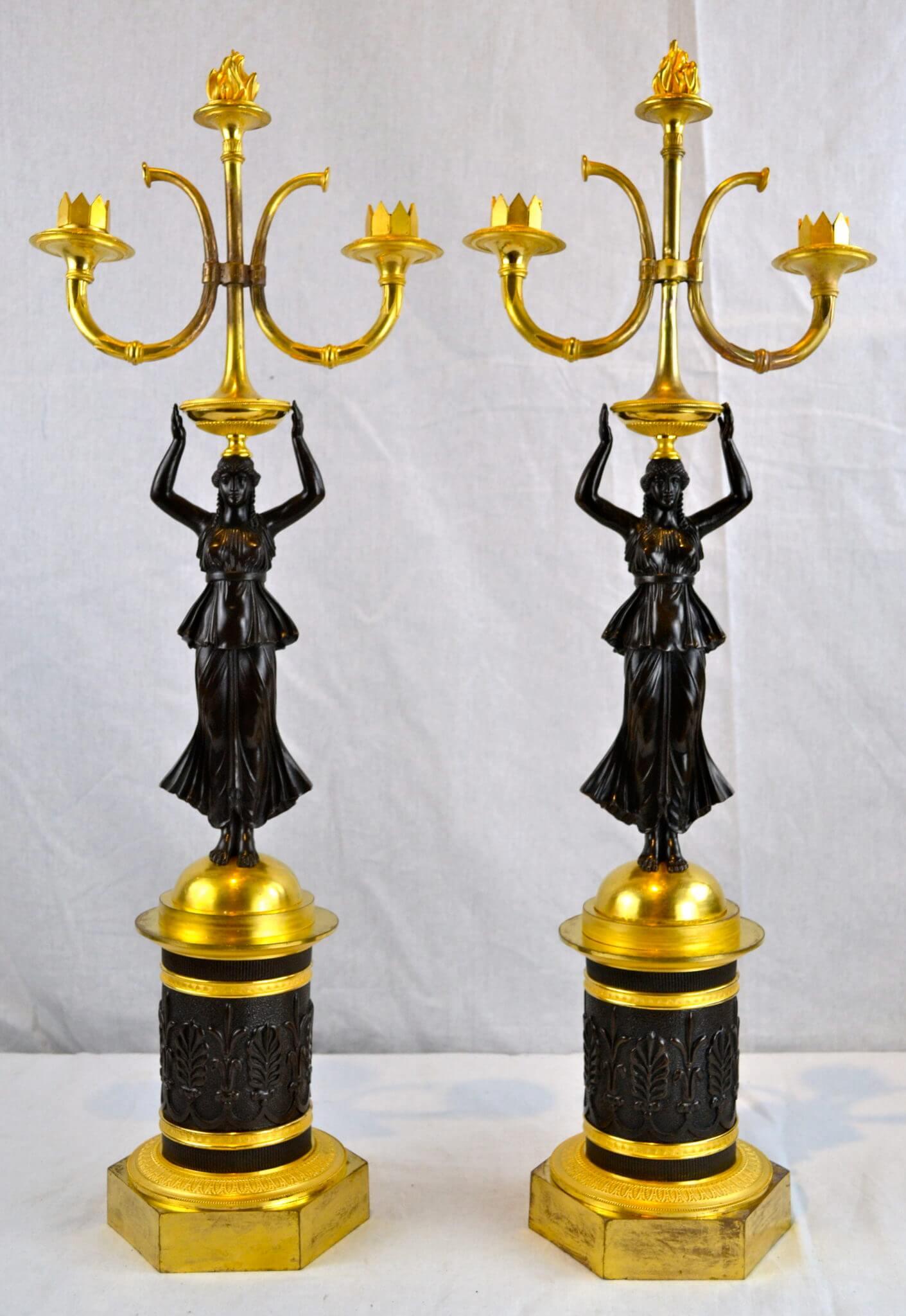 A pair of mid-19th century French Empire figural candelabra featuring a patinated bronze caryatid holding a gilt bronze urn, mounted with a torch atop her head. The whole raised on a gilt and patinated bronze domed columnar support, accented with
