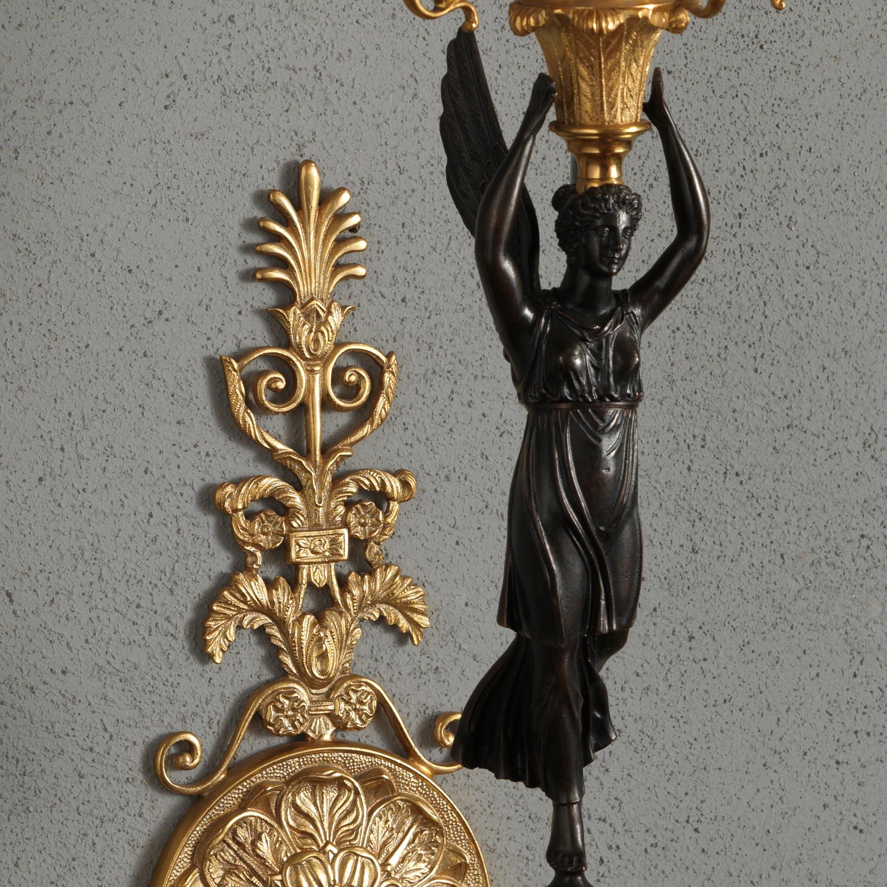 Dieser Wandleuchter im französischen Empire-Stil aus vergoldeter und brünierter Bronze ist ein elegantes Beispiel für eine figurale Wandleuchte, die eine geflügelte Victory zeigt, die auf einer kleinen runden Kugel aus vergoldeter Bronze sitzt. Die