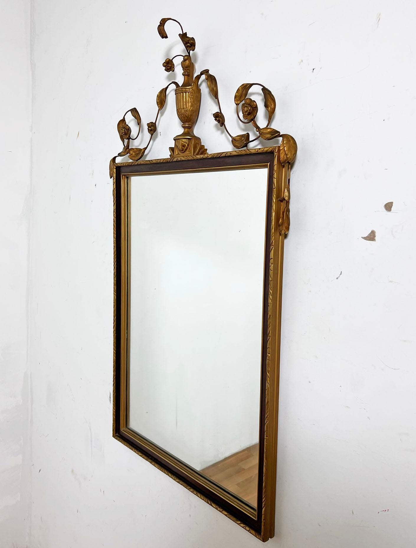 French empire style wall mirror featuring a classic motif of gilt leaf and flower vining emanating from a center urn, circa 1960s.