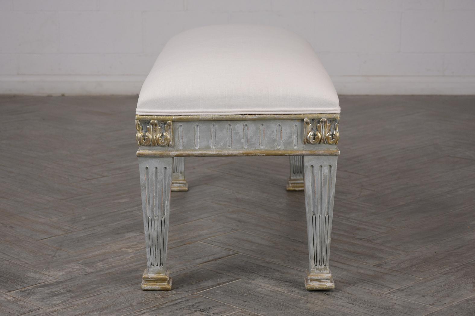 This 1900s Antique French Empire-style bench features a carved wood frame finished in a gray and white color combination with silver accents. The carved frame features architectural elements such as a frieze border and fluted legs with pedestal