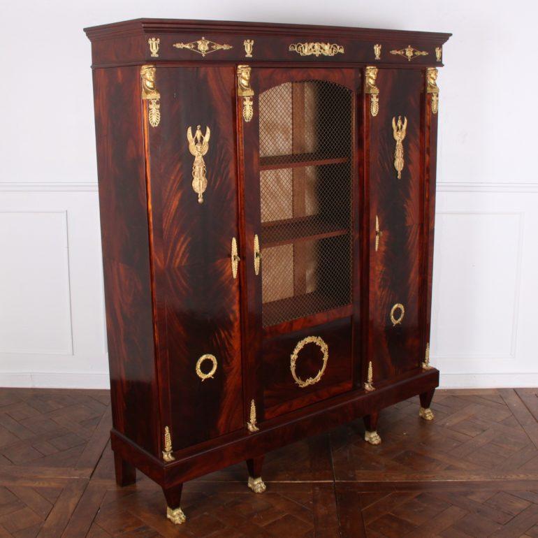 French Empire-style mahogany three-door bookcase, the two side-doors with flame mahogany and gilt bronze mounts and flanking a larger centre door with metal mesh front and lower ormolu ‘wreath’ detail. The piece features four pillaster columns