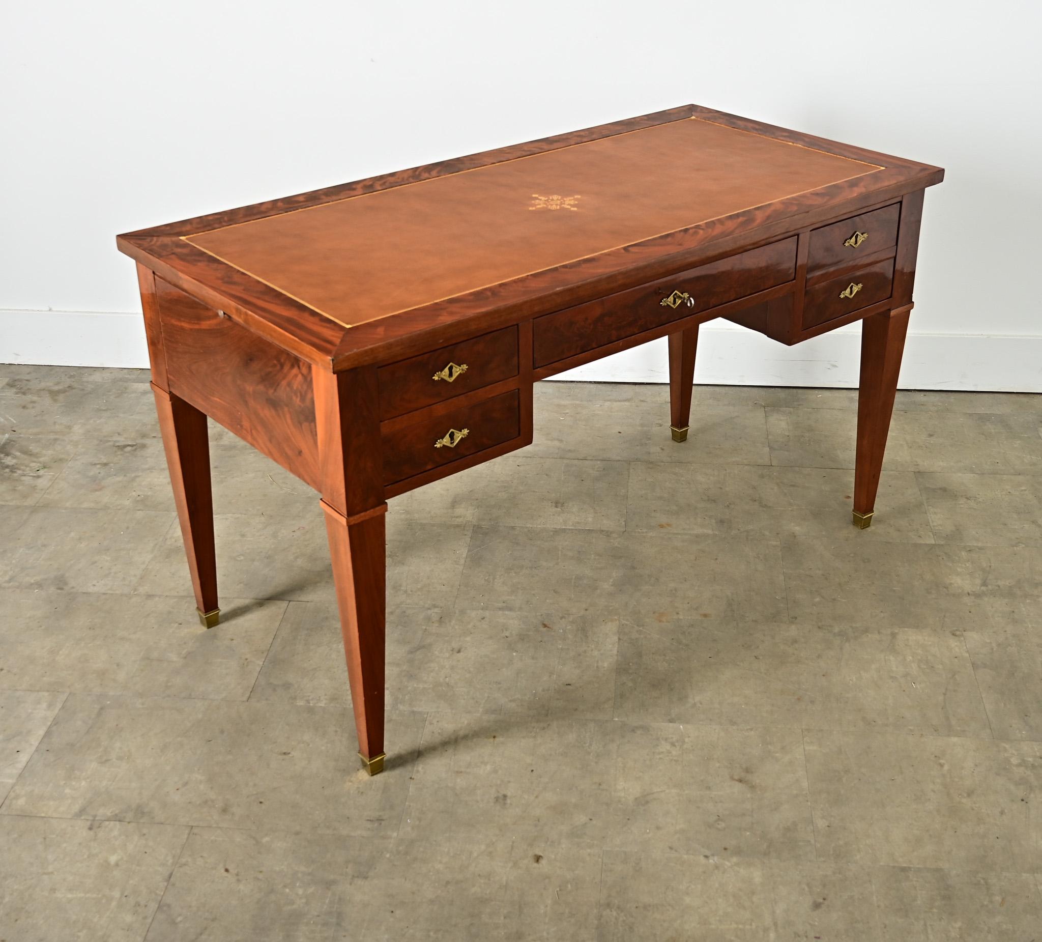 A French vintage 20th century leather-topped polished mahogany writing desk, designed in the Directoire taste for refined proportions & materials, accented with Neoclassical symbolism. The wide, cognac leather surface is gently worn and outlined in
