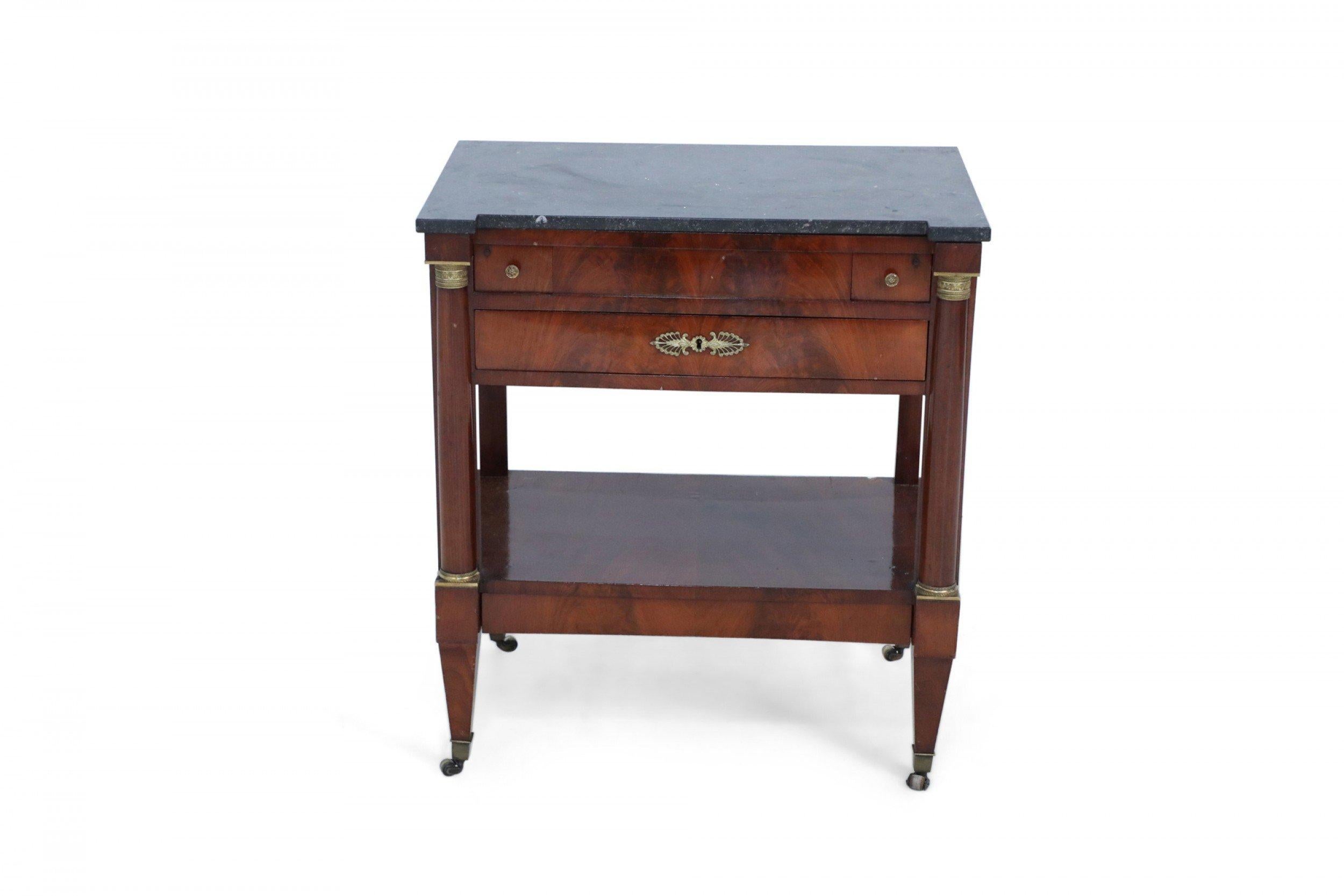 French Empire style mahogany two-tiered table with dark black and gray veined marble top with center ormolu embellishment above two small drawers and a center drawer supported on round legs with casters.