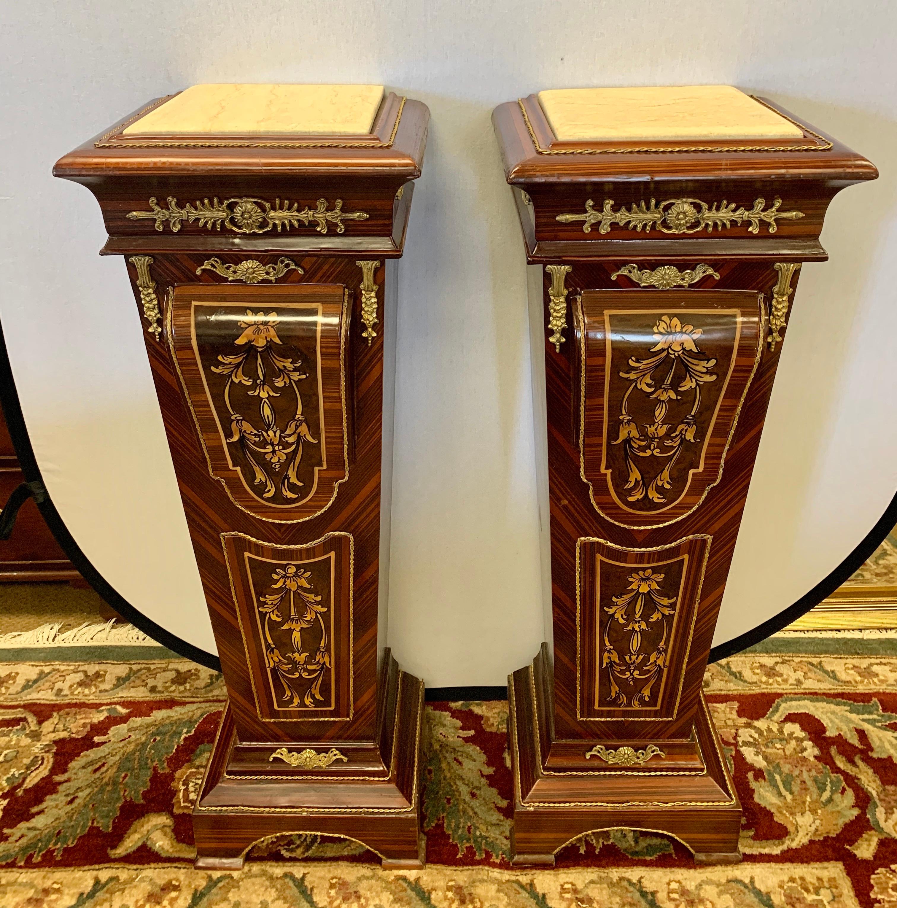 A pair of stunning French Empire style mahogany pedestals feature tall rectangular bodies, which taper towards their bases and are topped with white marble slabs. They feature gilt bronze ormolu mounts and floral scrolled detail on the front and