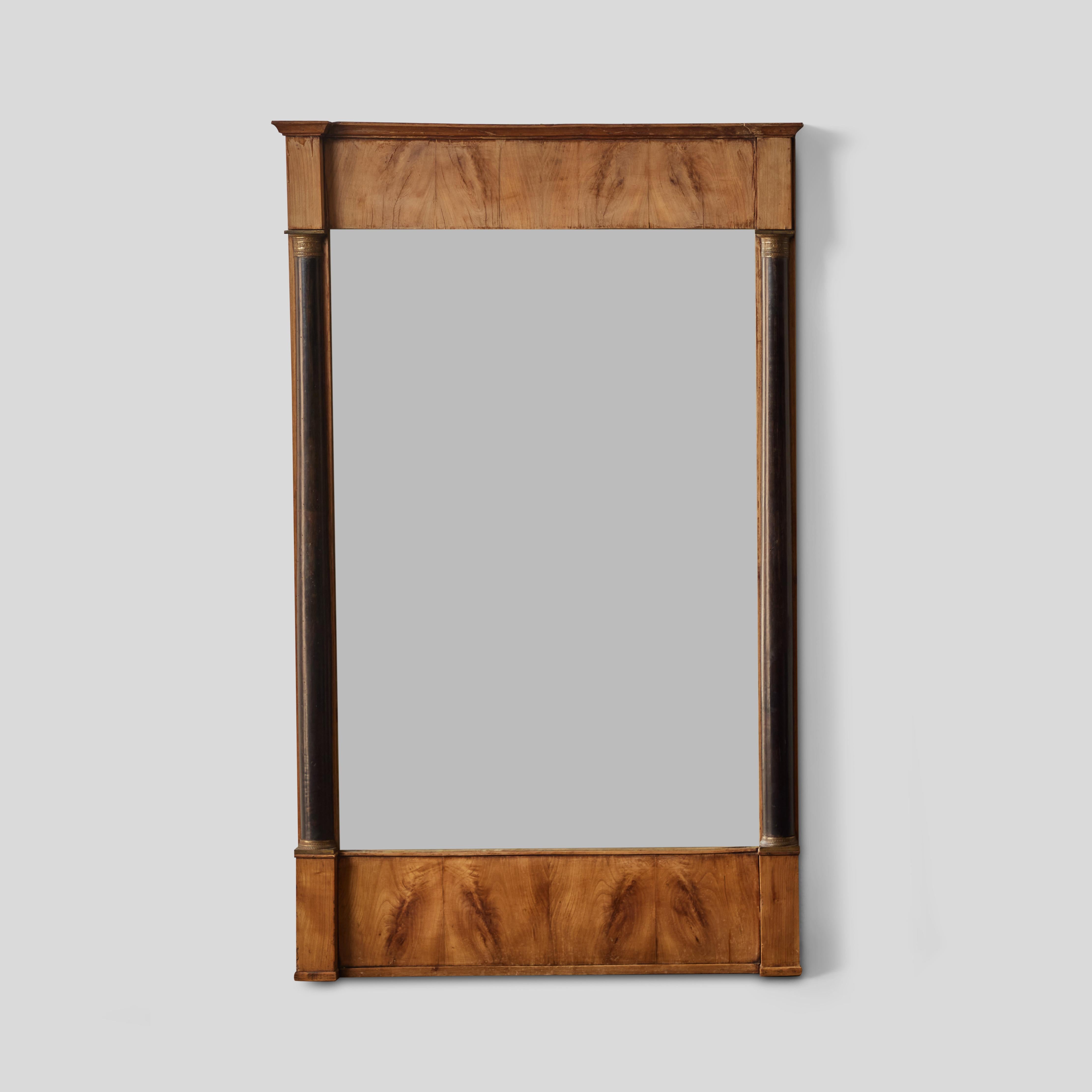 Questo specchio in stile French Empire è caratterizzato da eleganti colonne ioniche ebanizzate e da una cornice in legno burlato color miele. Il suo design incarna una sobria eleganza architettonica con una sensibilità proto-moderna, combinando
