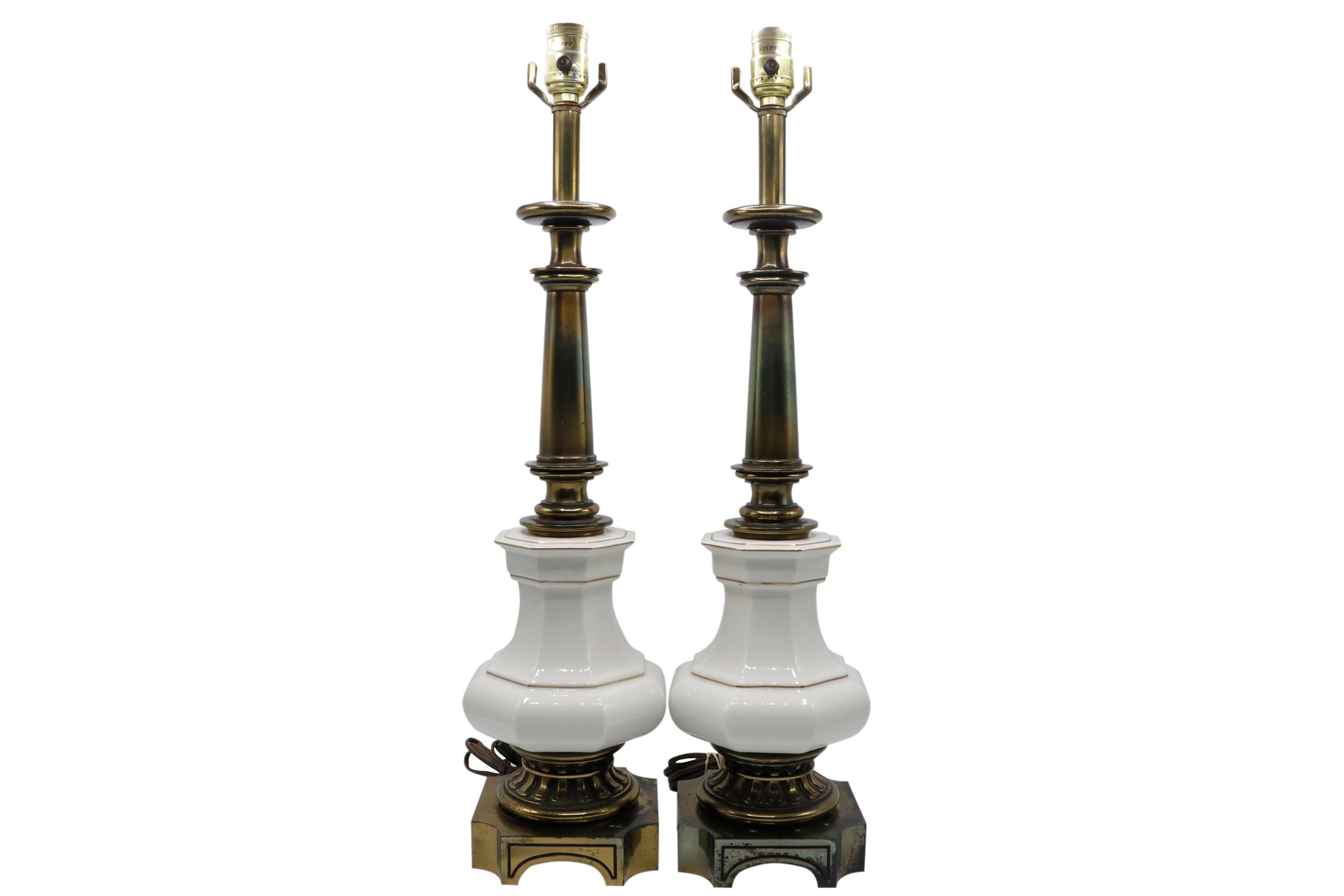 A pair of French Empire style table lamps made by Stiffel. Brass columns are turned with large ceramic fonts in white trimmed in gold. Bases are decorated with dentils on a square base with concave corners. Dimensions per lamp.