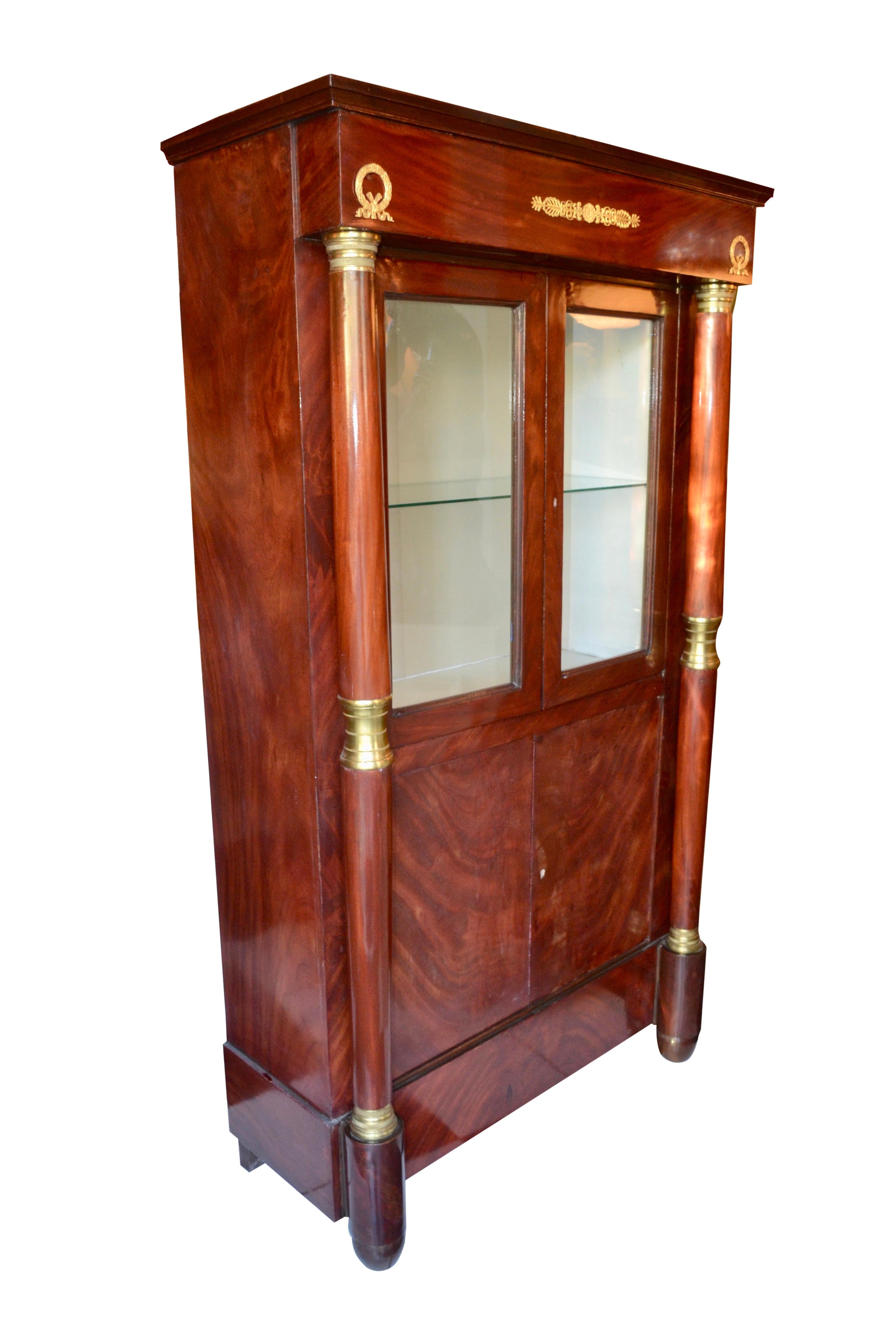A mahogany cabinet decorated with gilded metal mounts constructed in two sections, the upper section with two glazed doors flanked by mahogany columns opens to reveal a fabric lined interior with one glass shelf, the lower section with two book