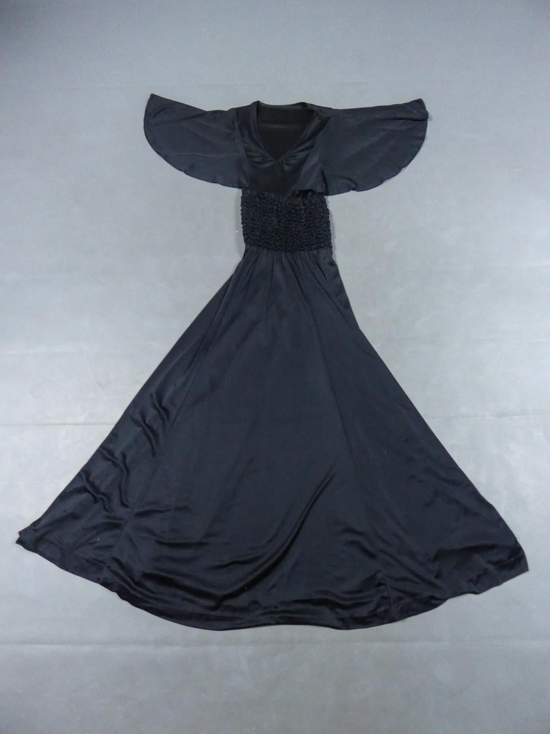 Alrededor de 1990
Francia

Vestido de noche largo de punto de seda negra strech de los años 90. Corte ceñido a la altura del corpiño que se abre desde la cintura dejando una falda fluida y suelta. Escote fruncido en V, mangas murciélago y cintura