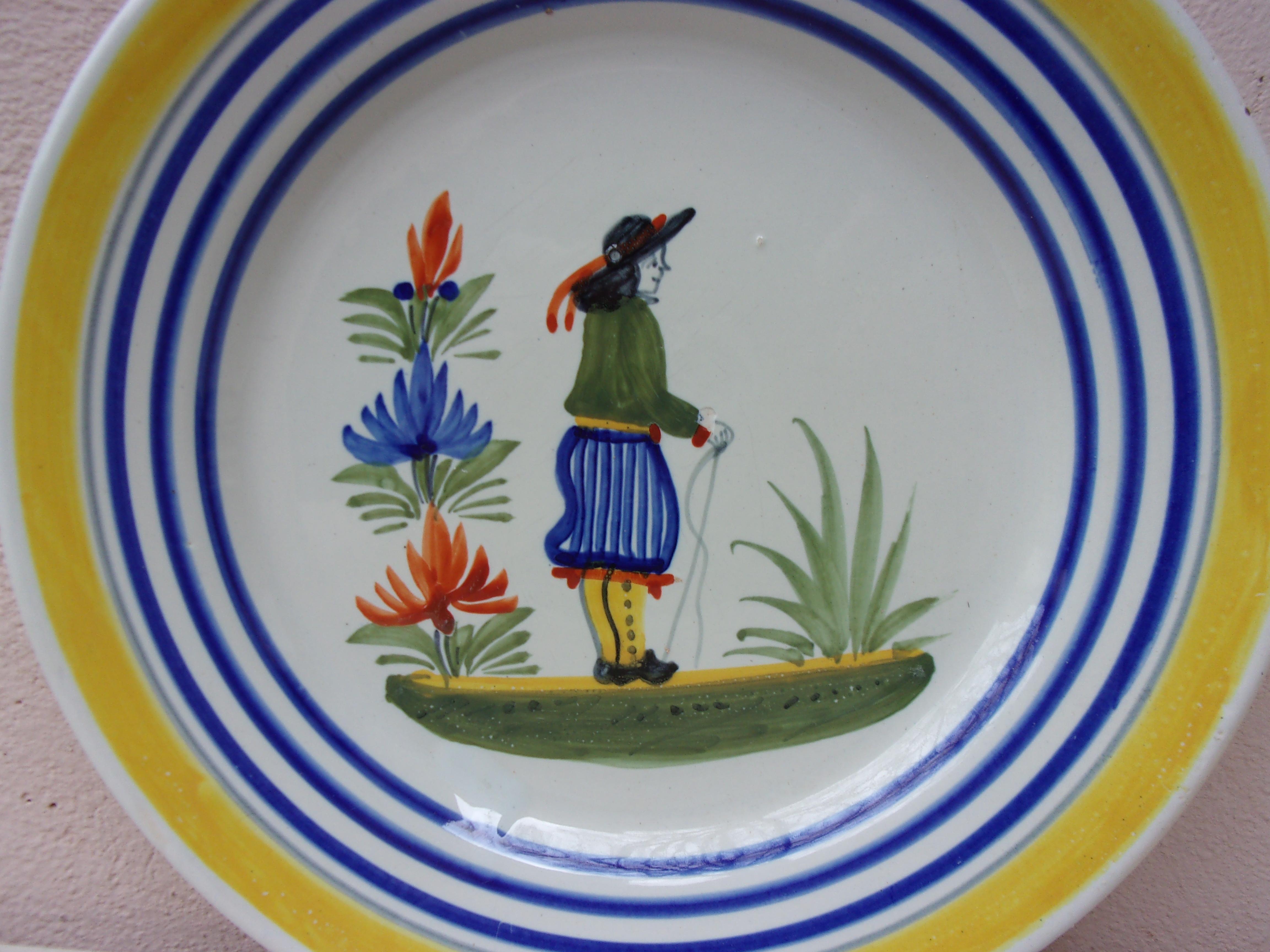 Assiette en faïence française représentant un fermier en costume avec des fleurs, signée Henriot Quimper, vers 1950.
Bordure jaune colorée et lignes bleues.
 