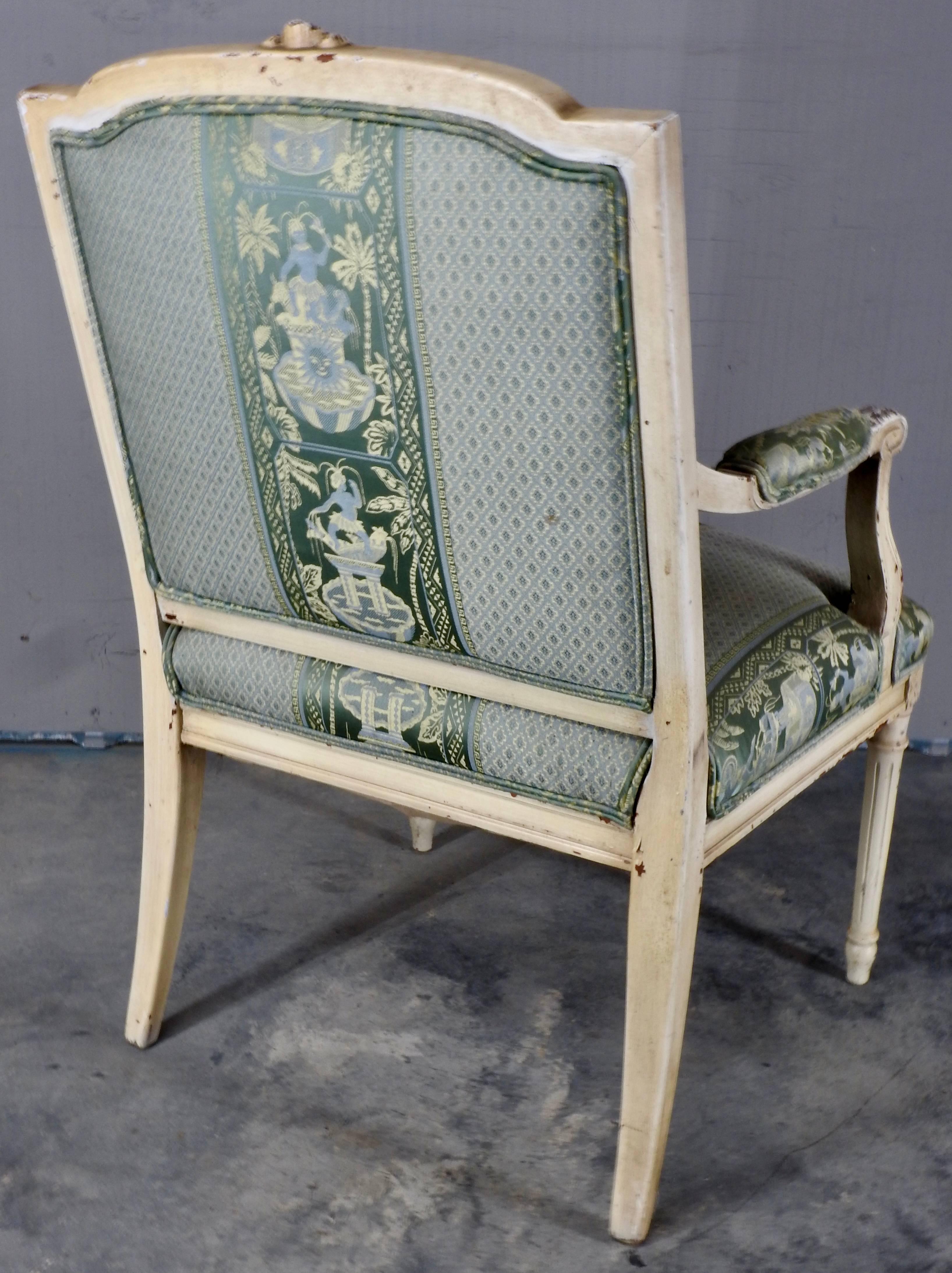 Distressed cream color paint highlights the details on this French fauteuil. The top of the chair features a bow with a trailing ribbon. The arms and legs have detailed carvings of floral and scrollwork. Soft green, blue and golden colors of the