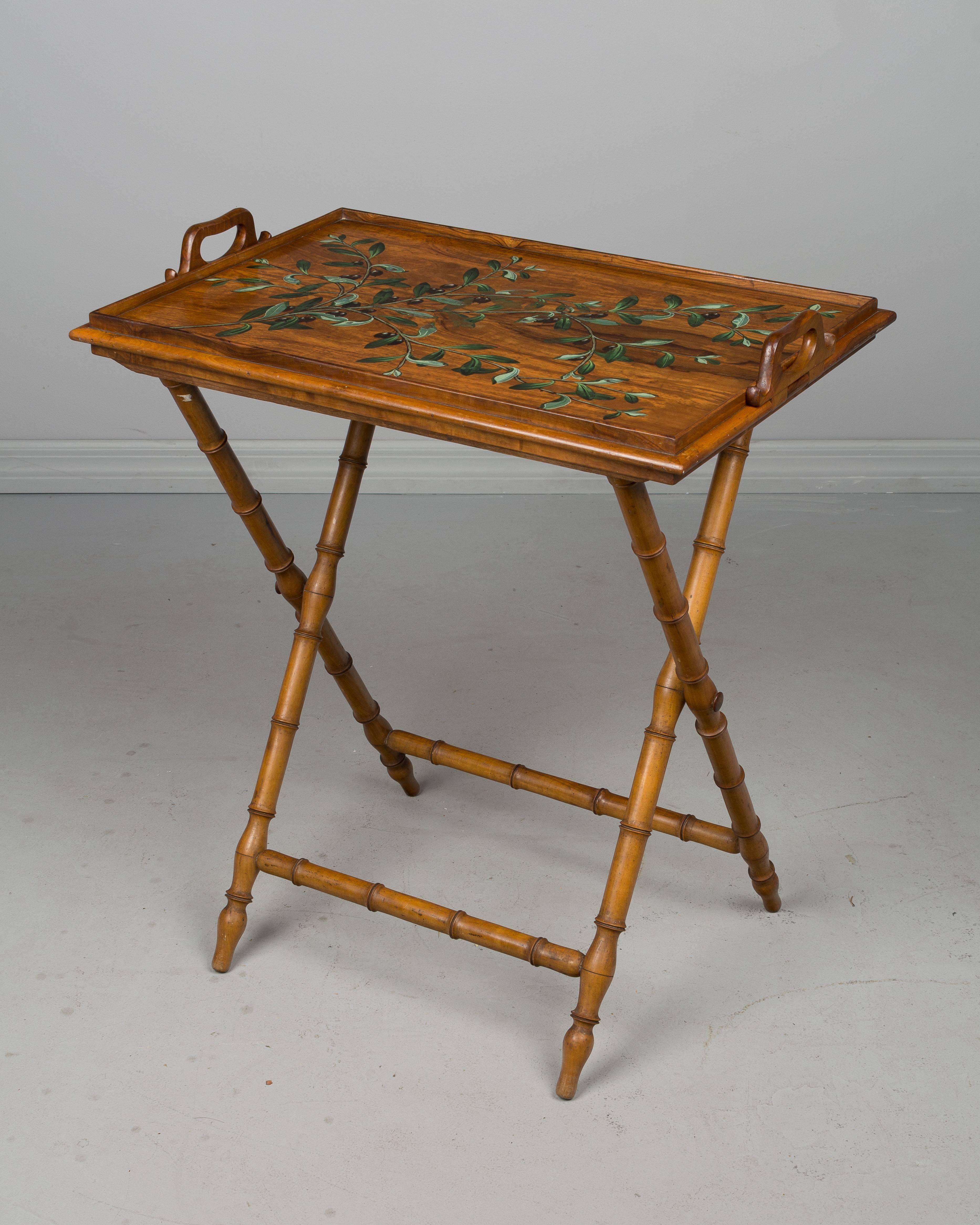 A French faux bamboo folding tray table with a base made of solid cherry and the reversible tray made of olive wood. One side of the tray is decorated with painted olive branches and the flip side has a red felted surface. The handles are hinged so