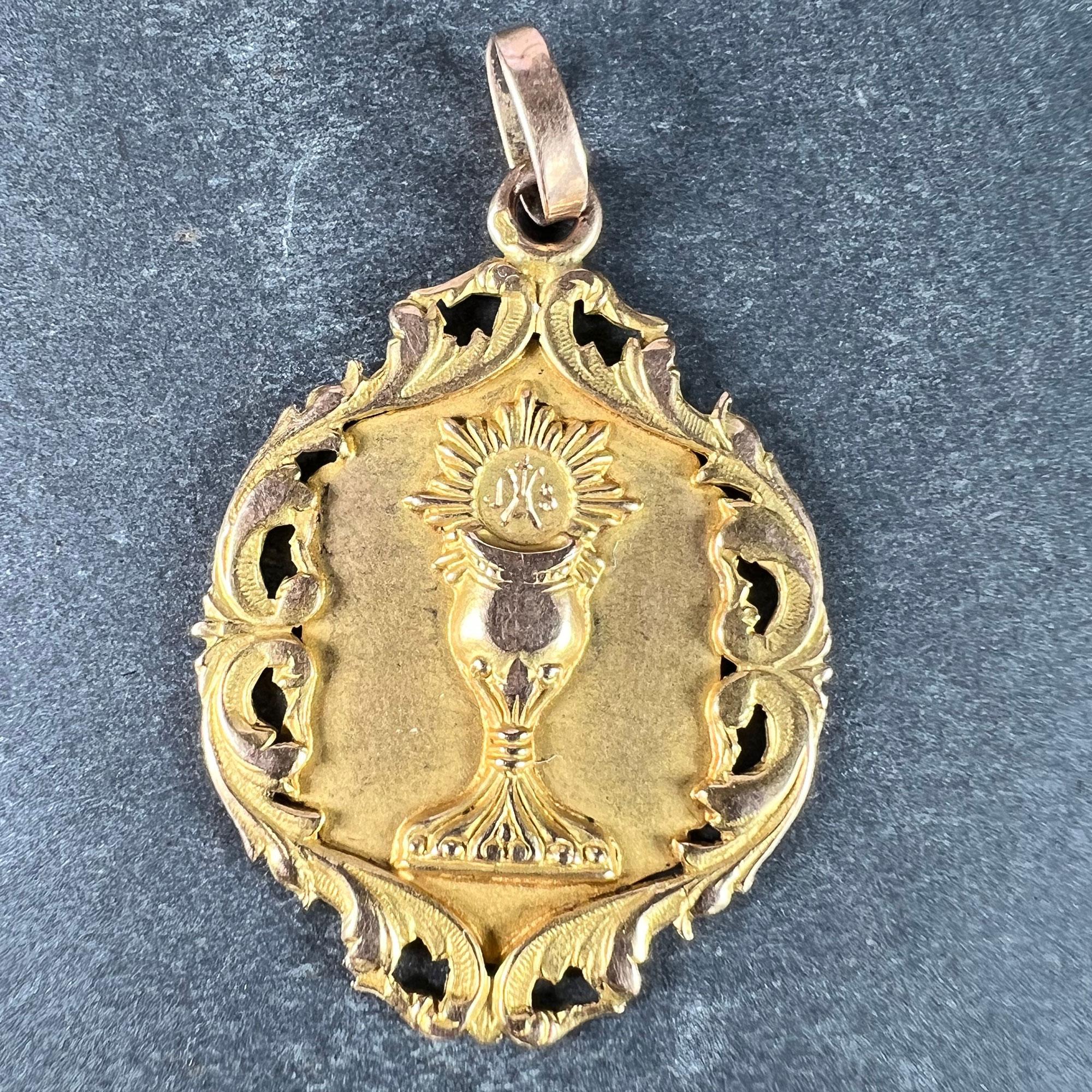 A French 18 karat (18K) yellow and rose gold charm pendant depicting a holy chalice or goblet with a communion wafer engraved IHS above, radiating light to celebrate a First Communion. The edges of the medal are scolled and foliate with pierced
