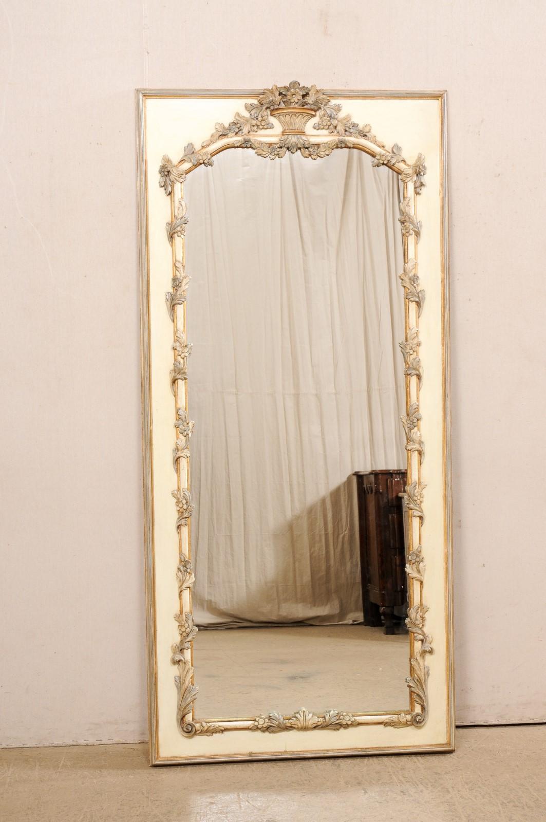 A tall French carved and painted wood mirror from the Mid-20th Century. This vintage mirror from France features a rectangular-shaped outer frame, with center mirror adorn about the inner surround with a carved upper crest topped with a bouquet of