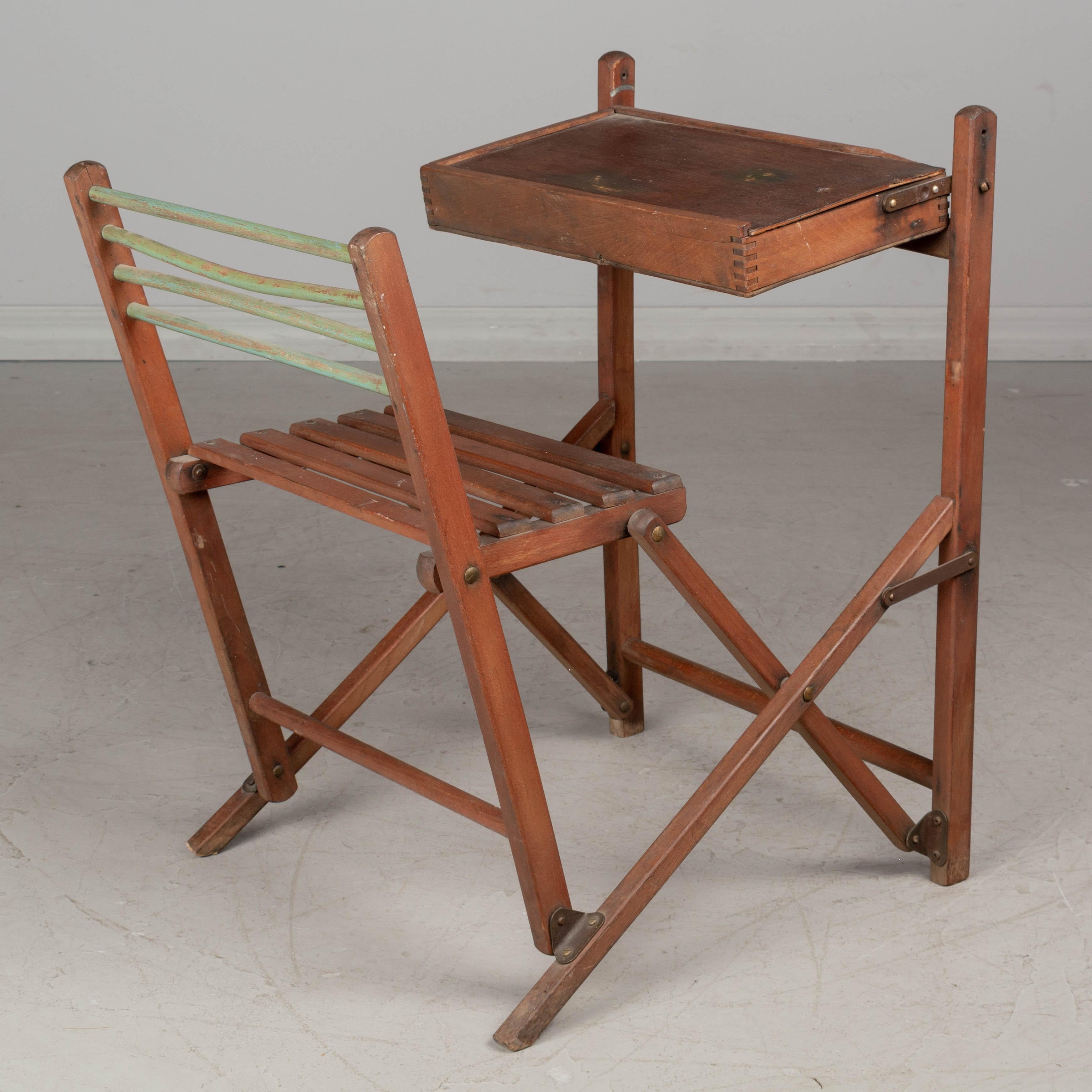 A French folding child's school desk and chair made of beechwood. The desktop has dovetailed corners and a sliding top for storage. The attached chair has a slat seat and metal rod back rest (one rode is slightly bent). Folds easily for storage.