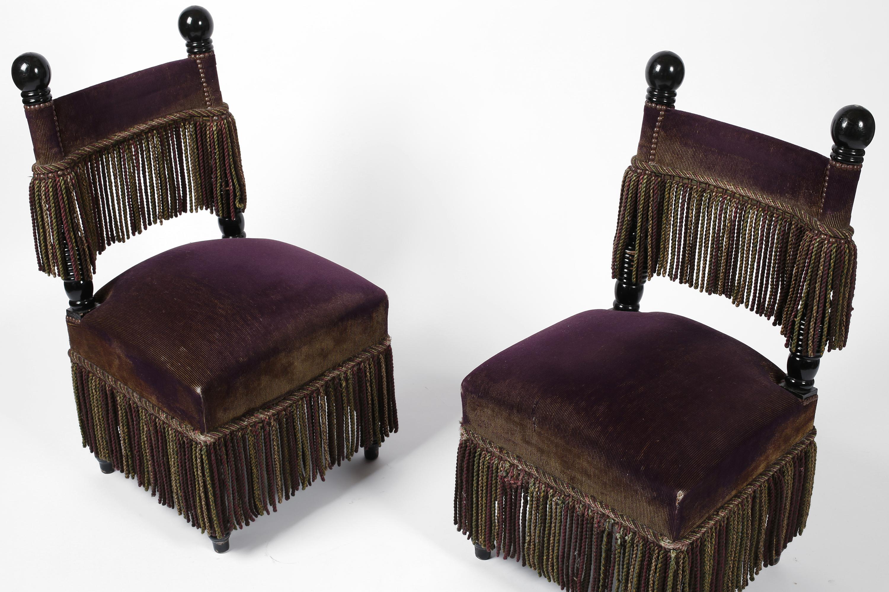 A duo of elaborate occasional chairs with turned and black lacquered timber frames, upholstered in their original plum corduroy velvet, adorned with brass studs, decorative trim and regal fringing. In the manner of Carlo Bugatti. 