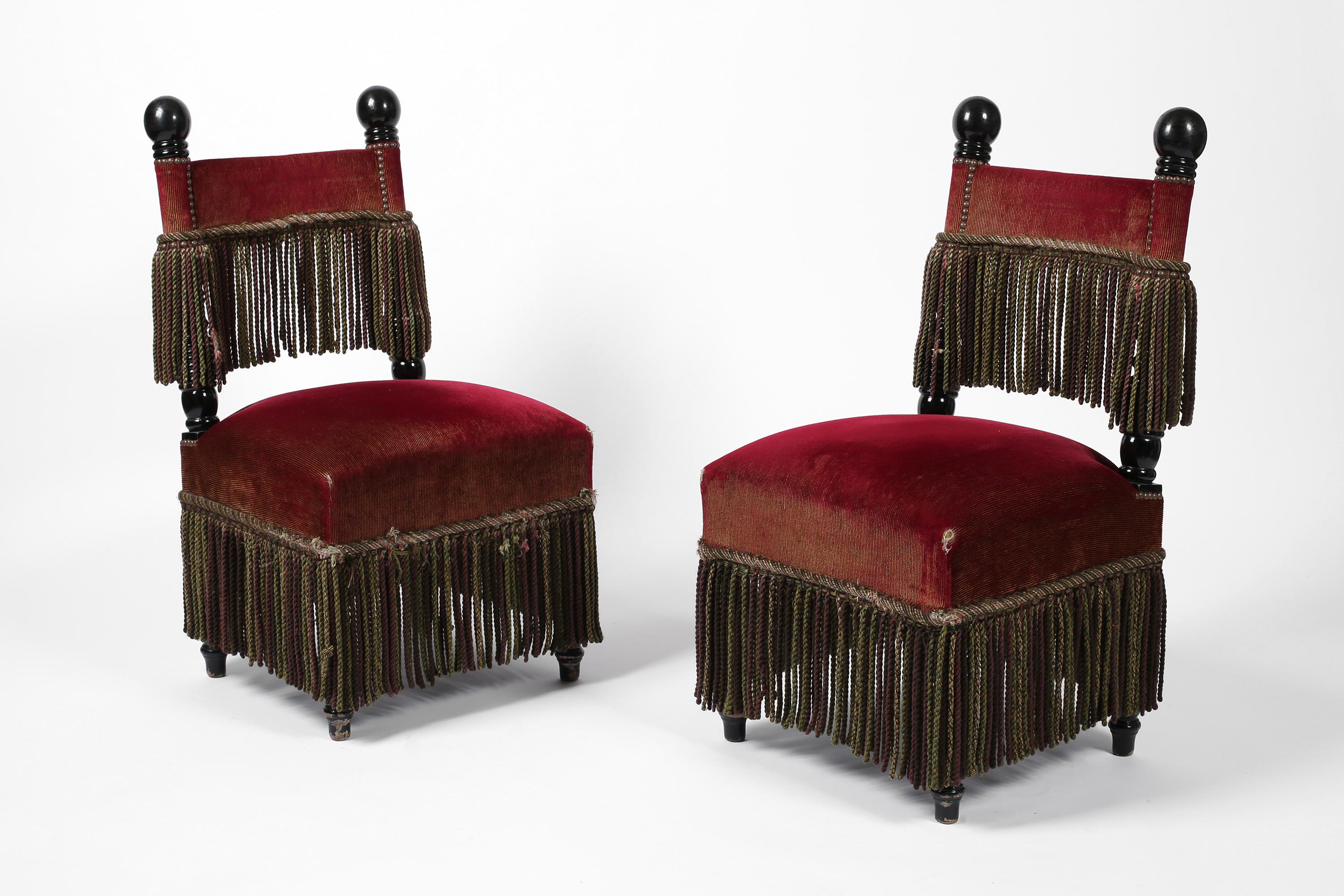 A duo of elaborate occasional chairs with turned and black lacquered timber frames, upholstered in their original ruby corduroy velvet, adorned with brass studs, decorative trim and regal fringing. In the manner of Carlo Bugatti. 