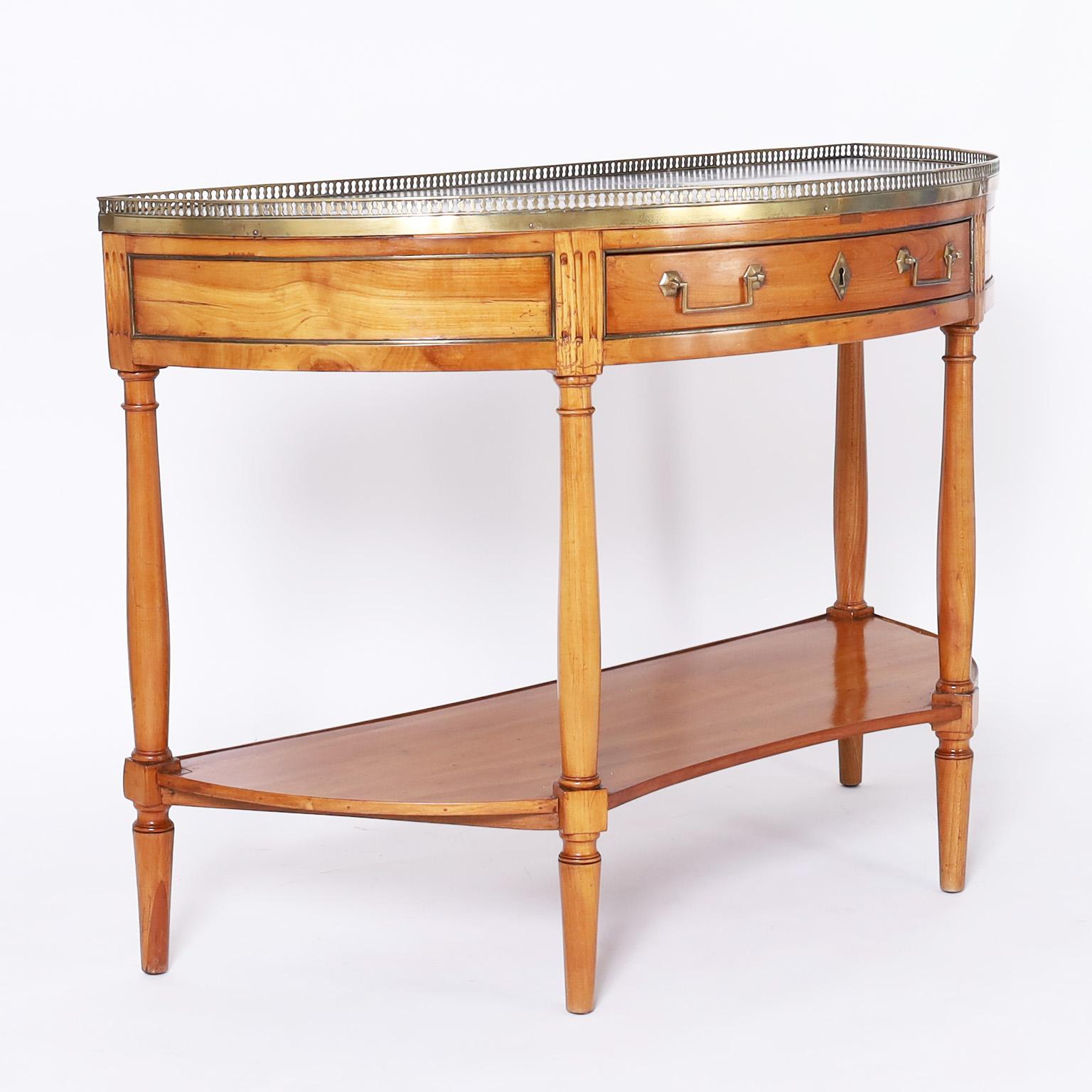 Antique French Directoire style server crafted in fruitwood with a demi-lune form having a black and white marble top with a brass gallery on a one drawer case with brass hardware on turned supports joined by a lower tier on tapered feet.
